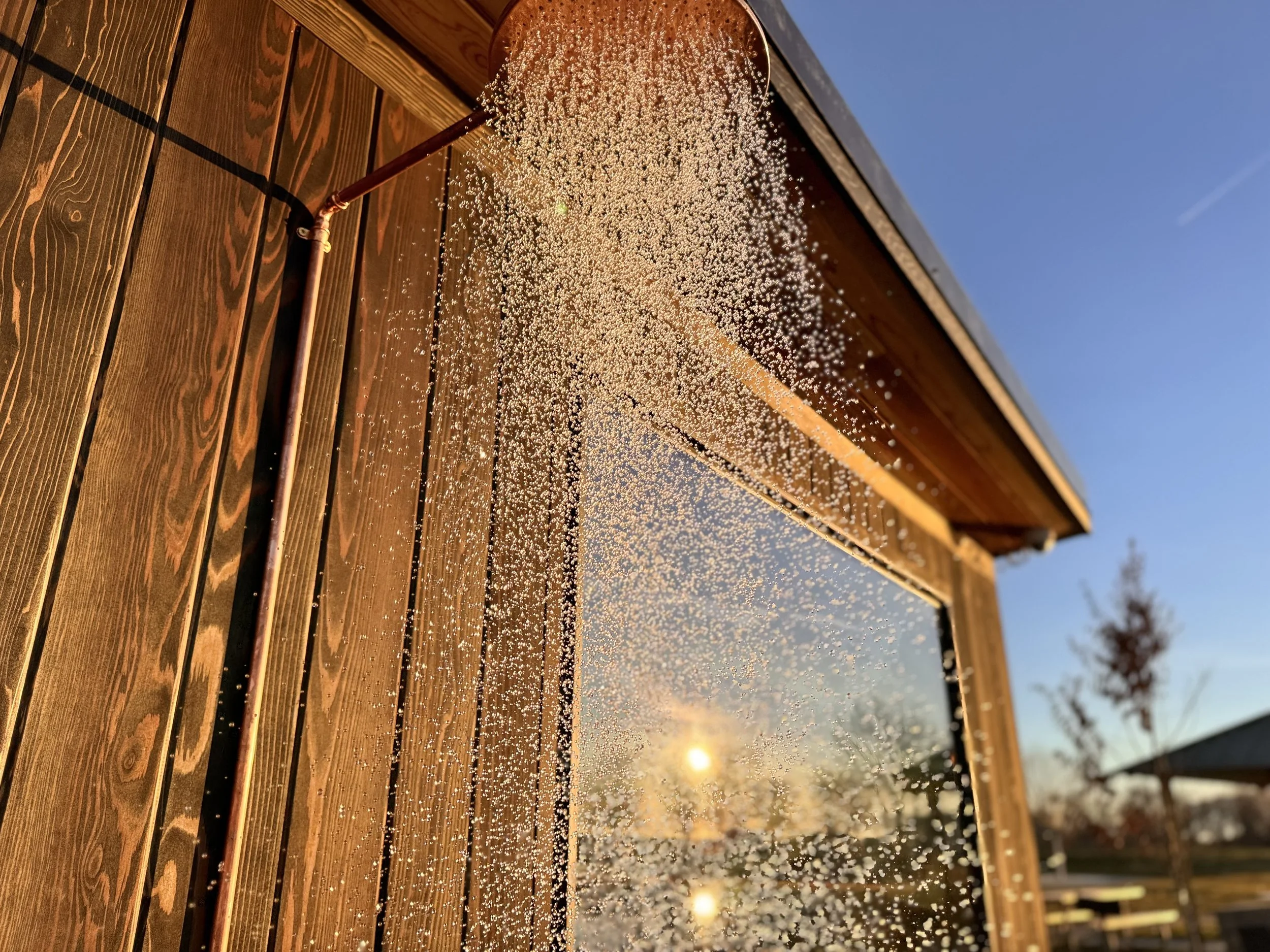 Sunshine reflecting off a rain-covered window next to a wooden wall. The sky is clear blue with a few clouds and a tree in the distance.
