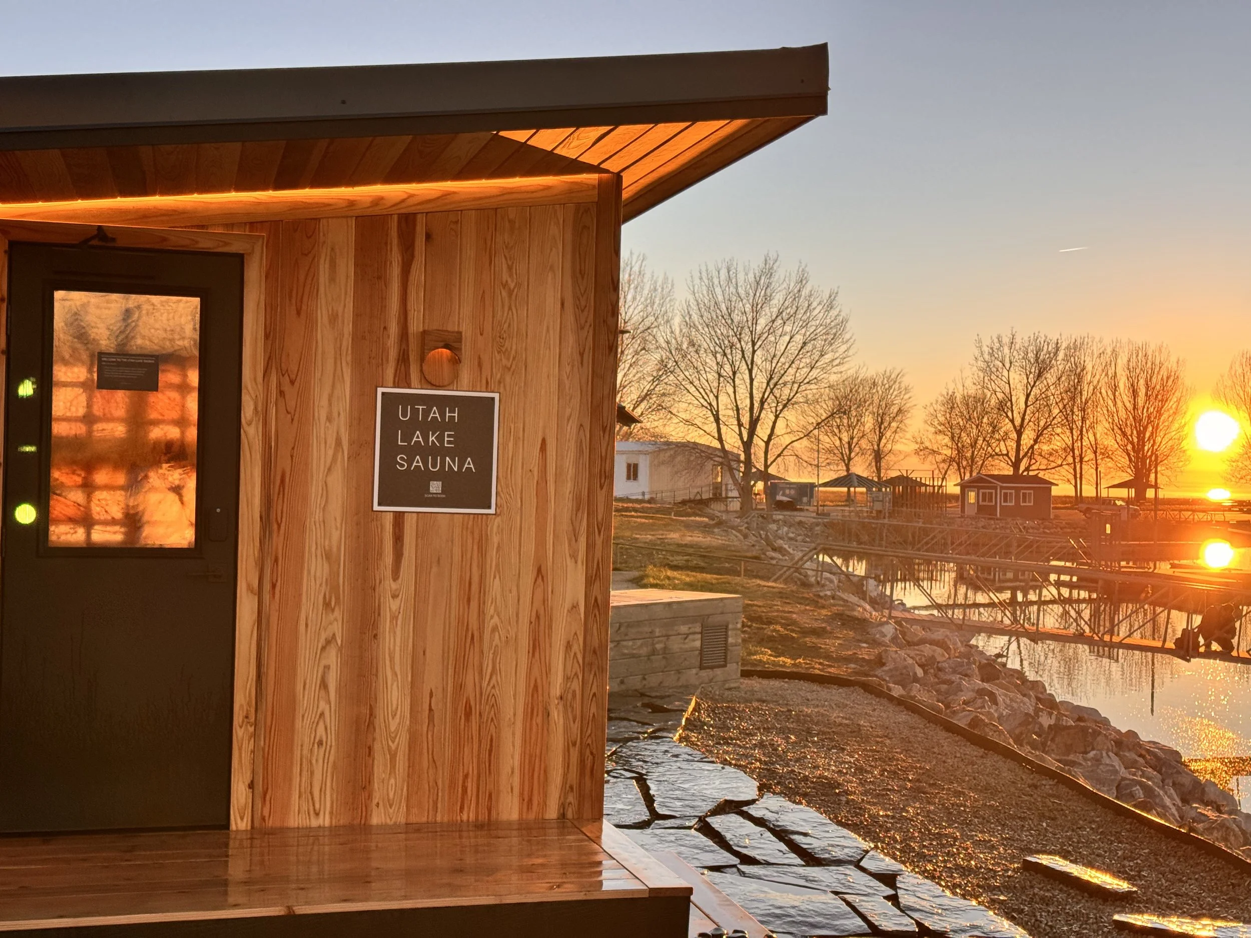Wooden building with a sign that reads 'Utah Lake Sauna' at sunset, overlooking a river with leafless trees and small buildings in the background.