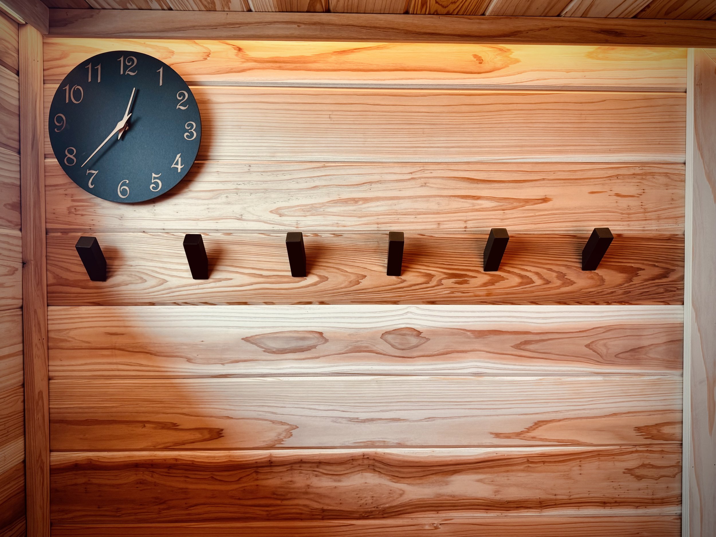 A blue clock showing 12:03, mounted on a wooden wall with five black hooks underneath.