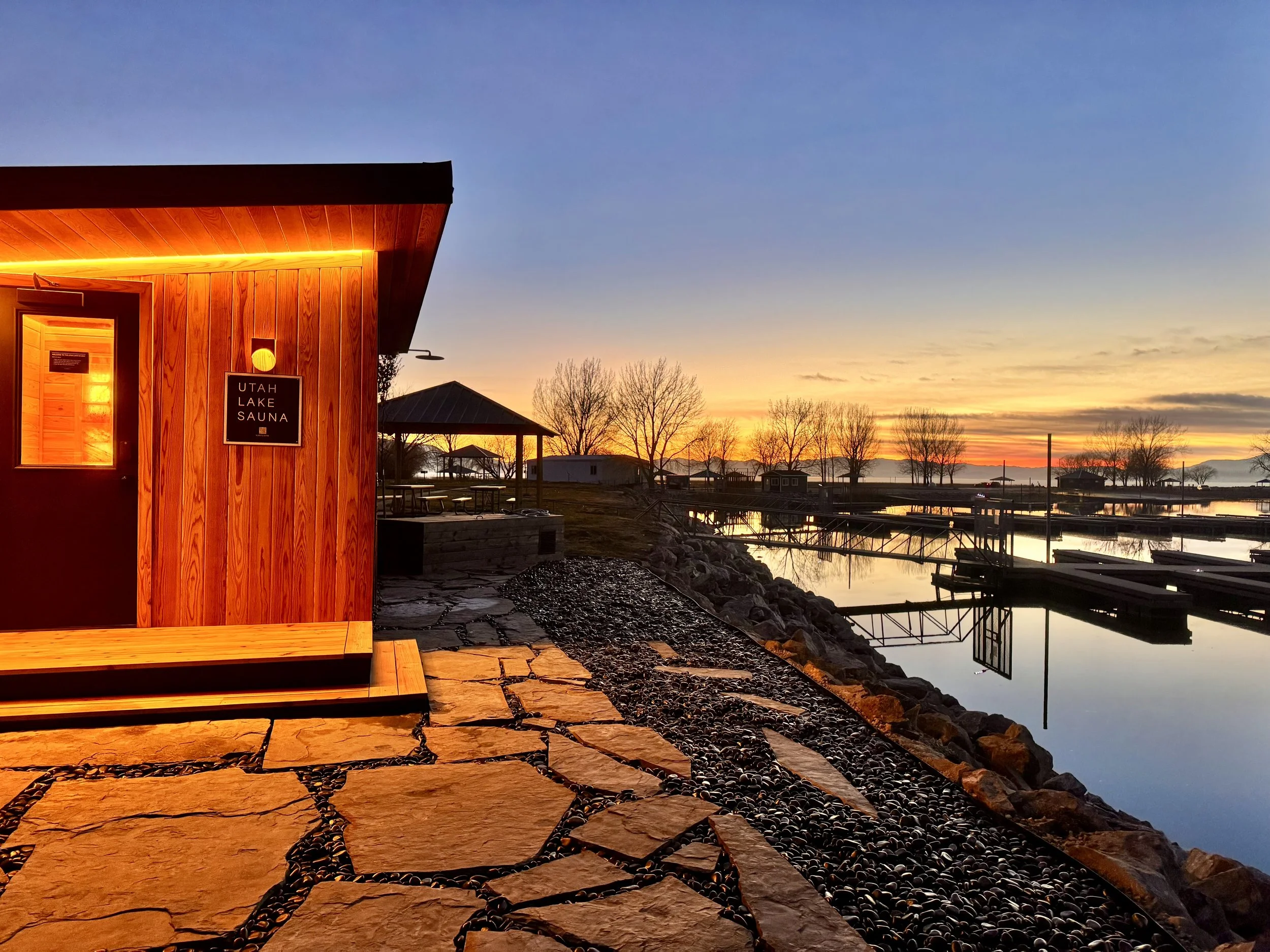 Sunset over a dock at Utah Lake with a wooden sauna building on the left and bare trees in the background.