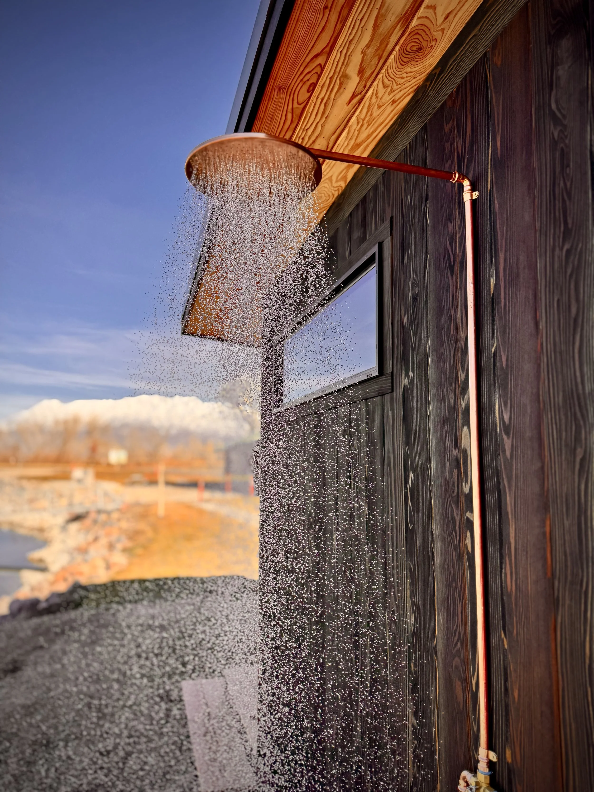 Outdoor copper showerhead releasing water on a wooden exterior wall with a small window and clear blue sky in the background.