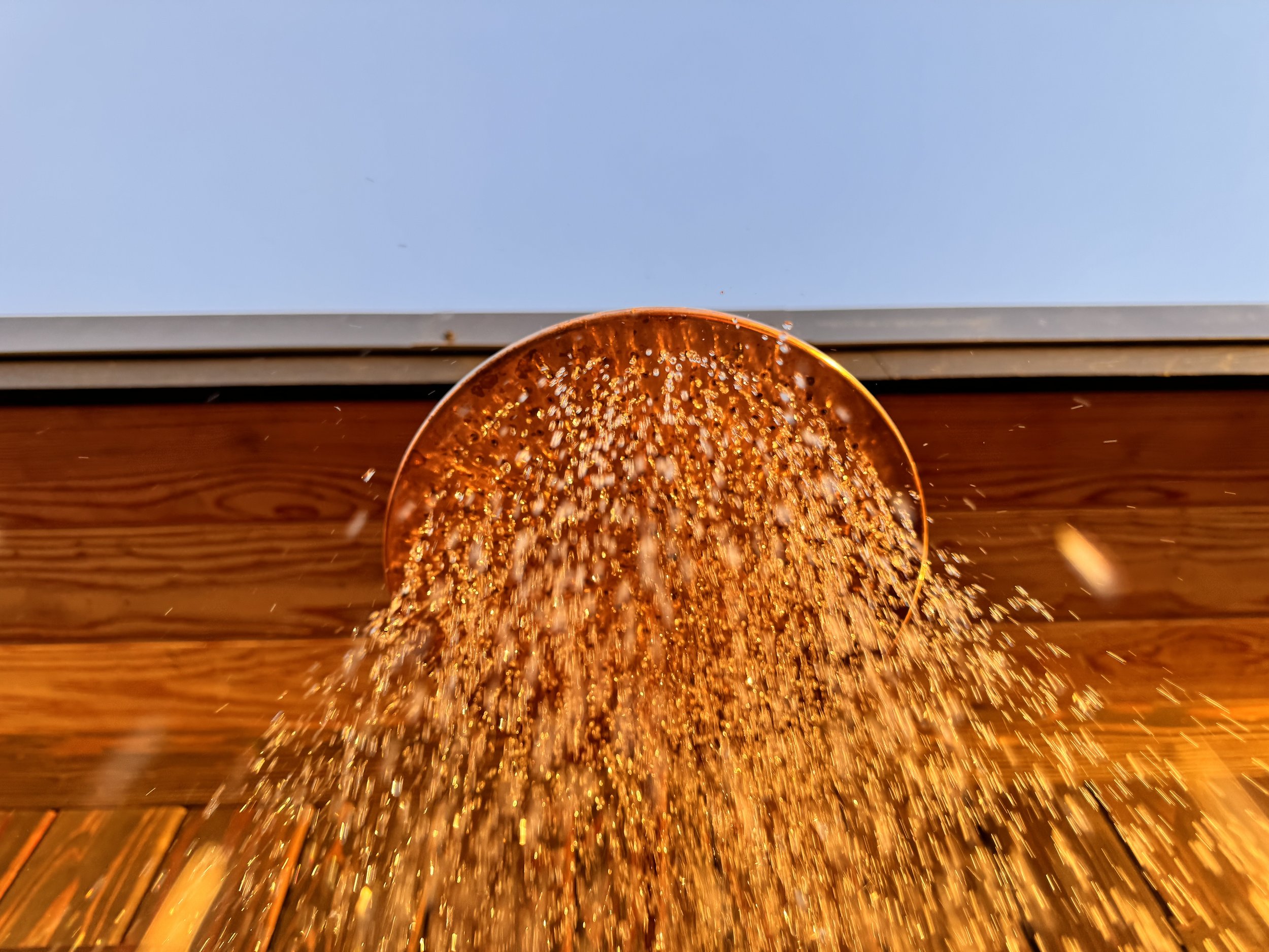 A copper cup tilted, pouring a stream of small particles onto a wooden surface outdoors.