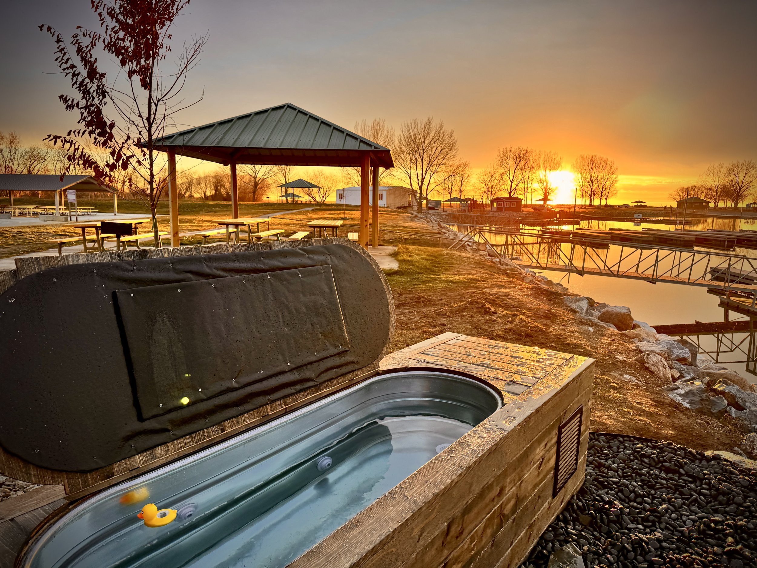 A hot tub with a rubber duck in a scenic outdoor setting during sunset, with picnic tables, gazebos, and a body of water in the background.
