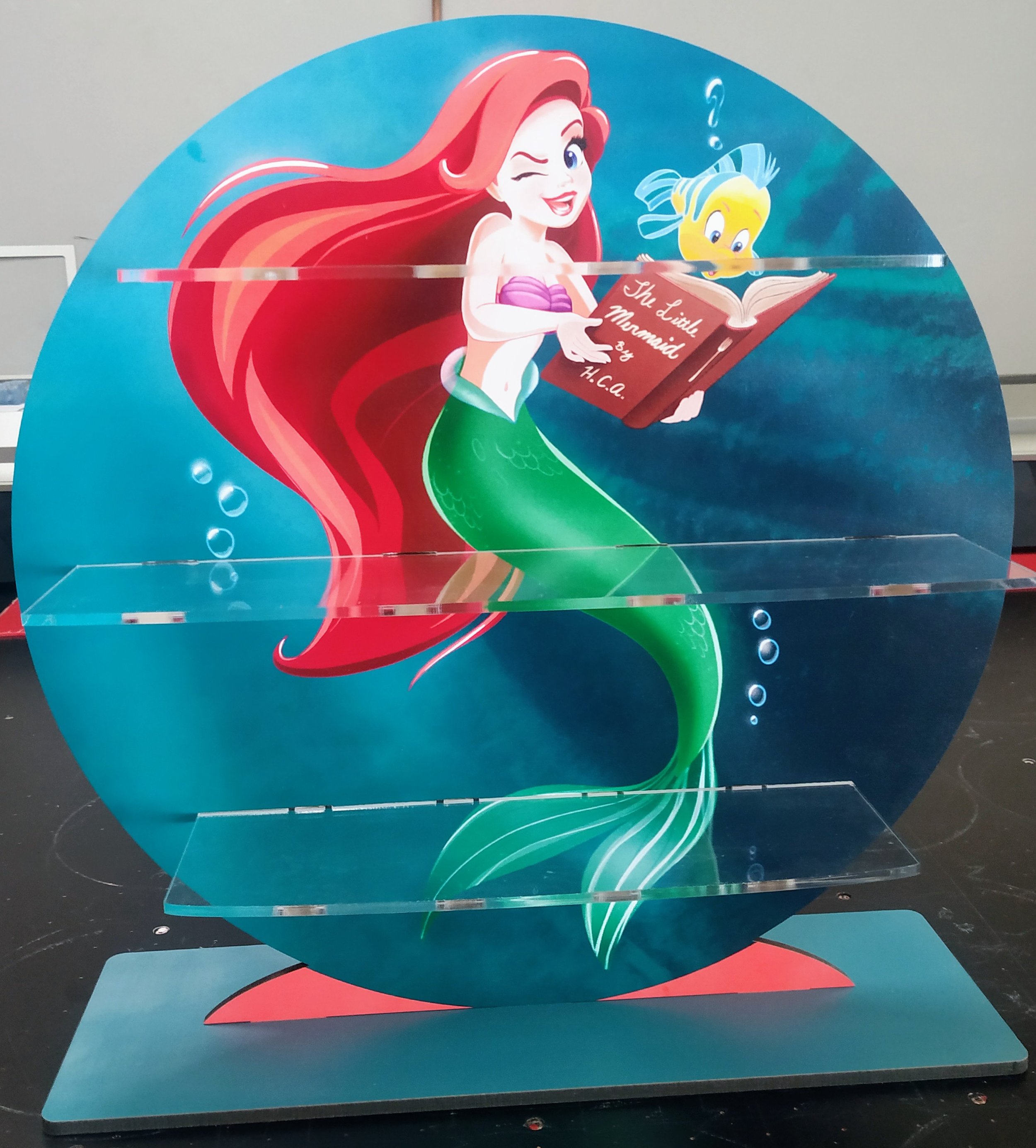 Decorative circular shelf featuring a colorful illustration of Ariel, the mermaid from Disney, holding a book titled "The Little Mermaid by H.C.," with Flounder, her fish friend, above the book. The shelf has three clear acrylic tiers.