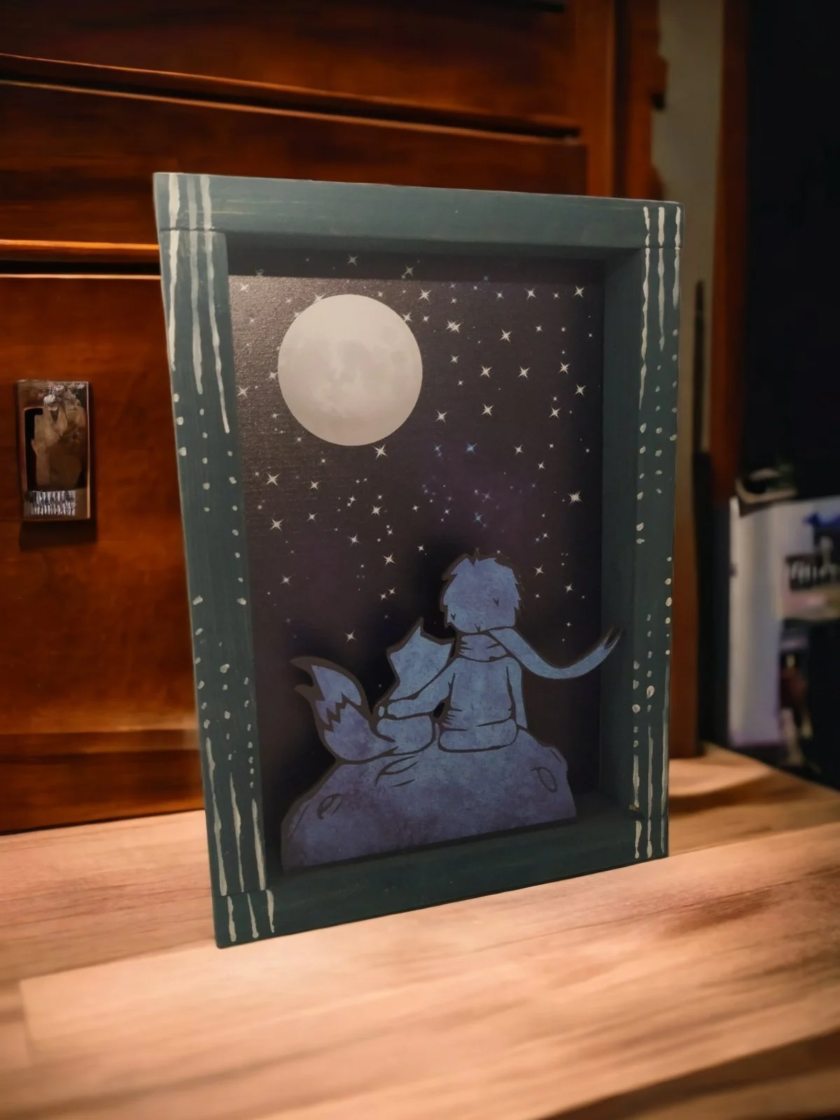 Decorative night scene with a cutout of a boy sitting on a rock, watching the full moon and stars.