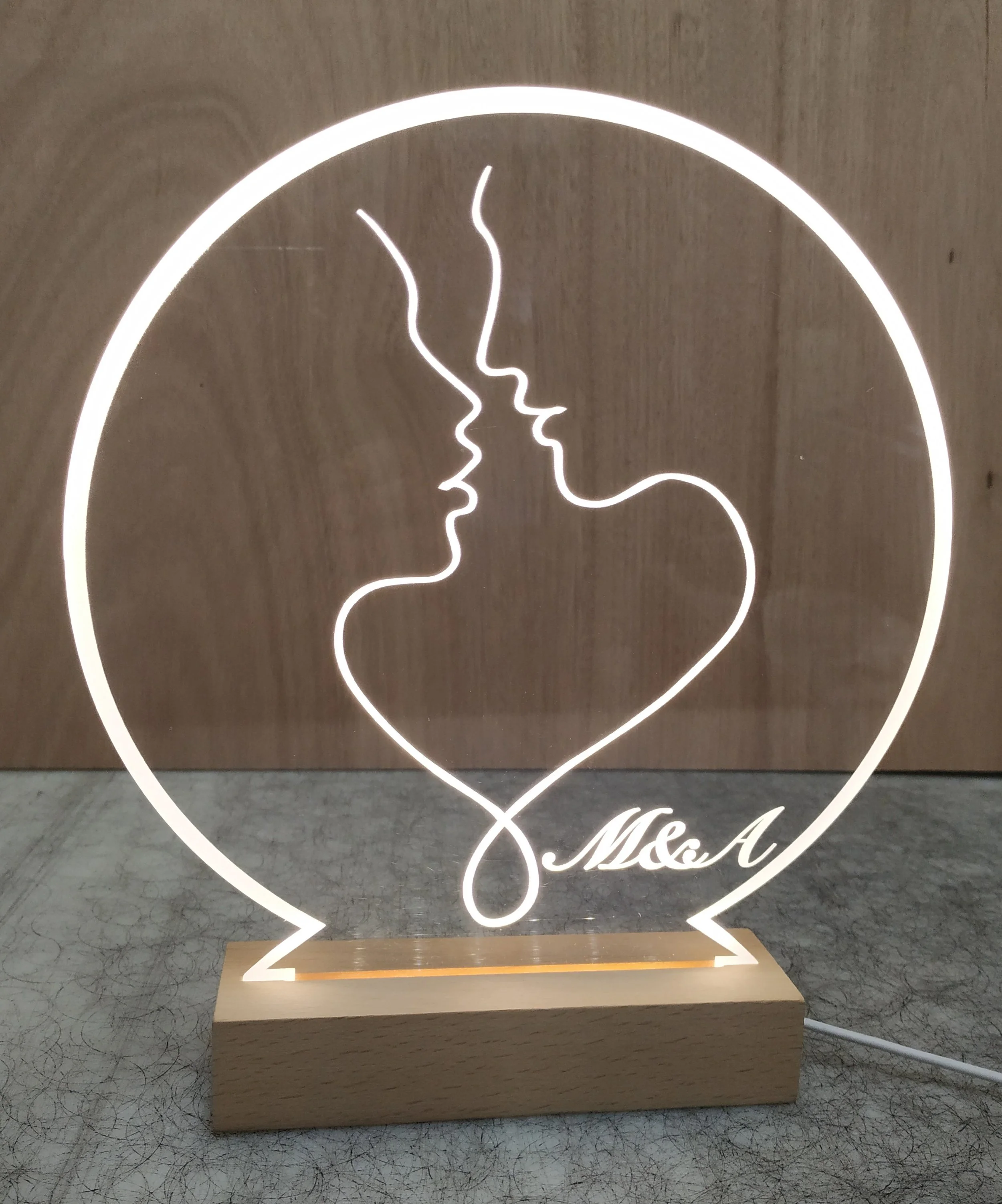 Neon light sign featuring two faces in profile, a man and a woman, with their foreheads and lips touching, forming a heart shape with the line. The sign is mounted on a wooden base with the inscription "Ms. A".