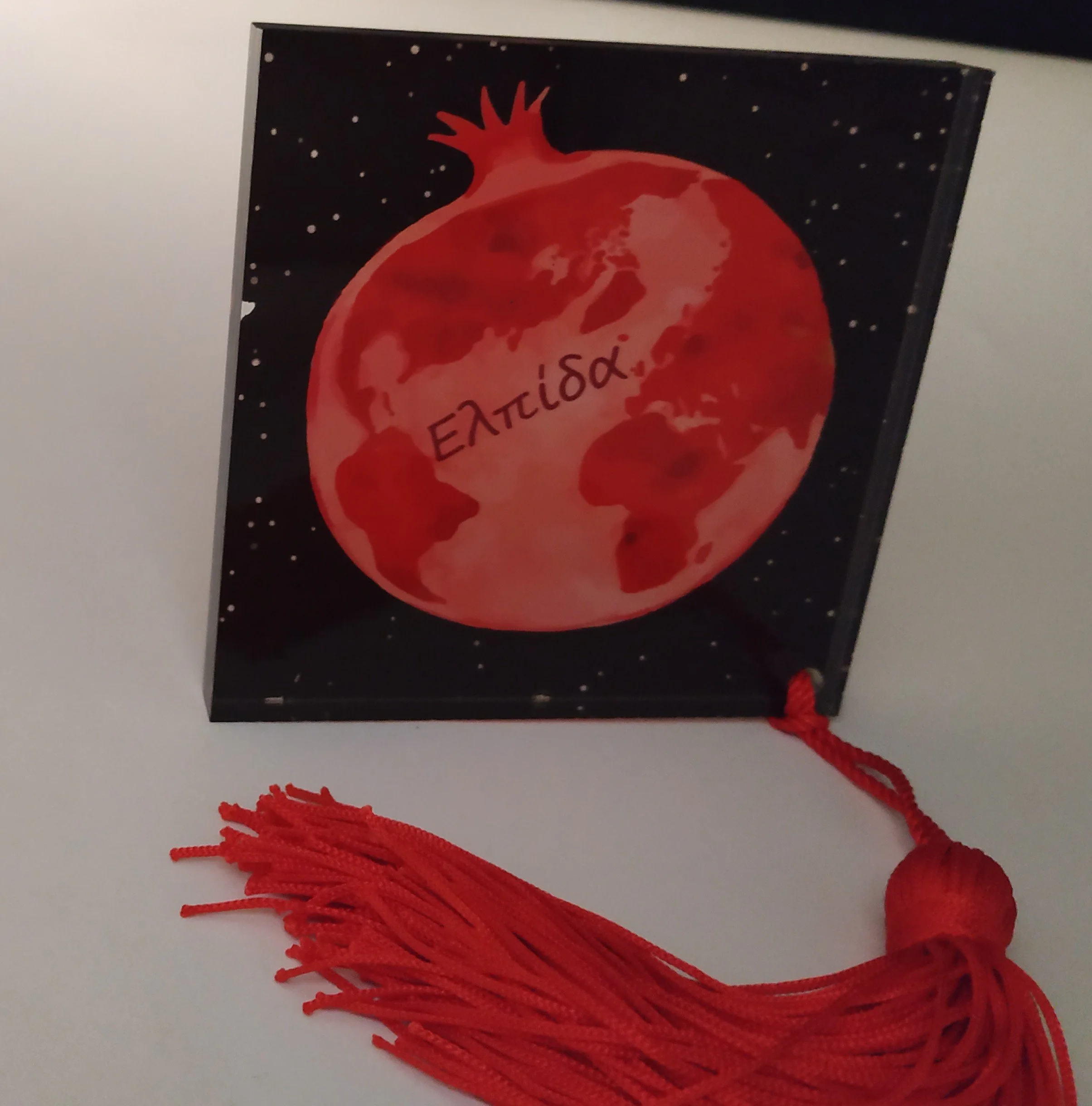 A graduation cap decorated with a red moon and world map design, with Greek letters, and a red tassel.