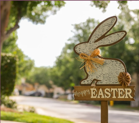 Easter bunny decoration with a sign that says 'Happy Easter' on a pole in a park with trees and a street in the background.
