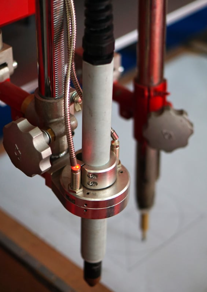 Close-up of a mechanical device with metallic pipes and tubes, featuring a sensor or laser component with a red light, connected to a support frame.