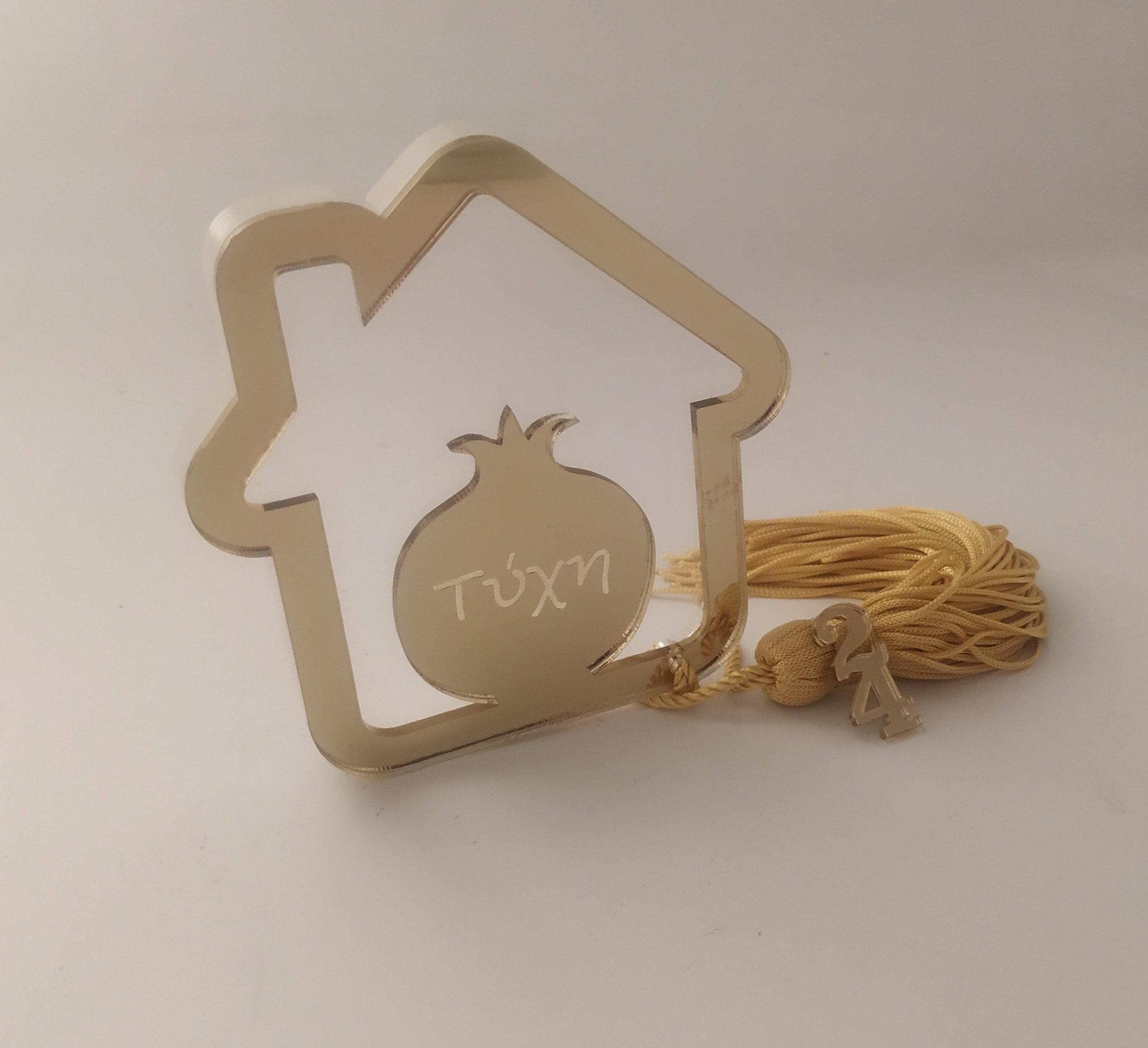 Gold ornament in the shape of a house with a pomegranate cutout in the middle, inscribed with the word 'Túxm'. Attached to a yellow tassel with a small charm.