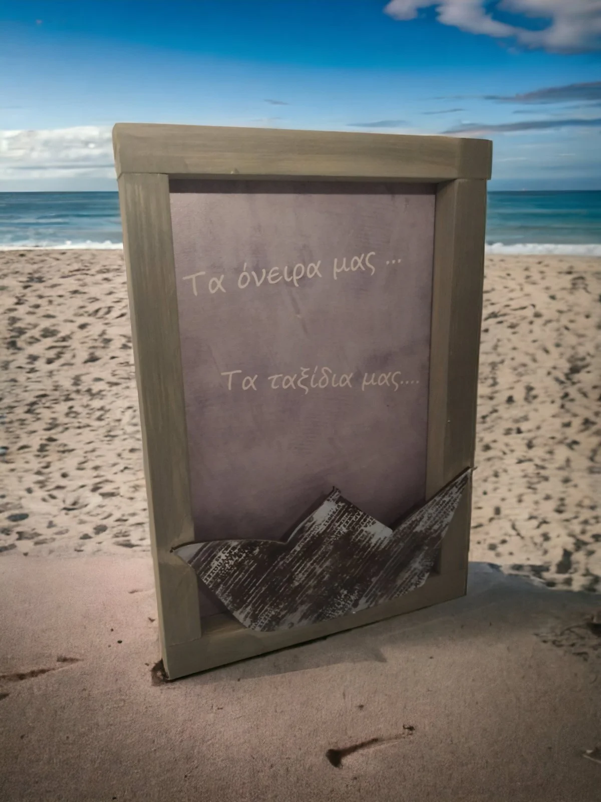 A decorative sign on the beach with Greek text, featuring a paper boat and a scenic ocean background with cloudy sky.