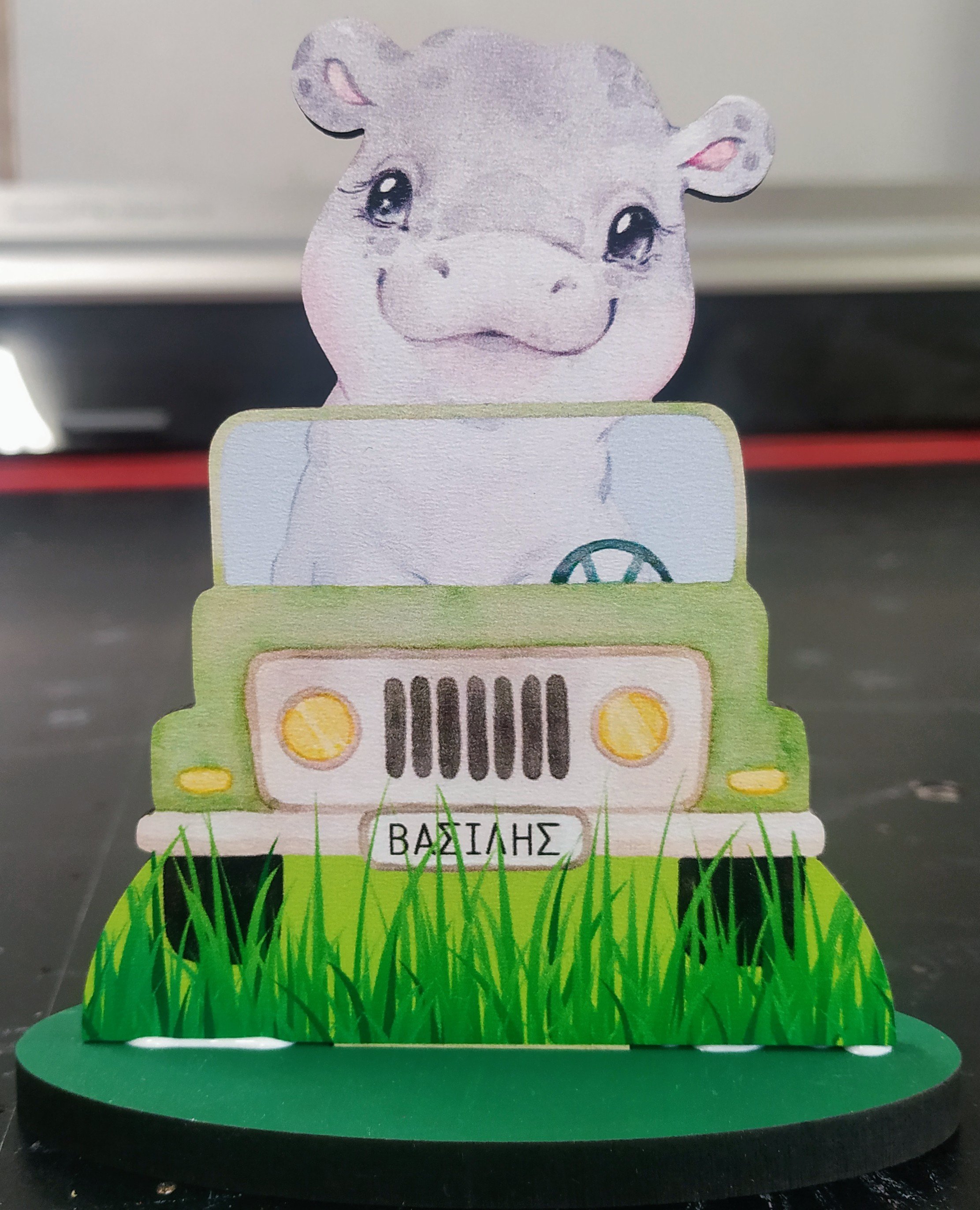 A paper cutout of a smiling hippopotamus driving a green vehicle with grass at the bottom, and the name 'ΒΑΣΙΛΗΣ' on the front.