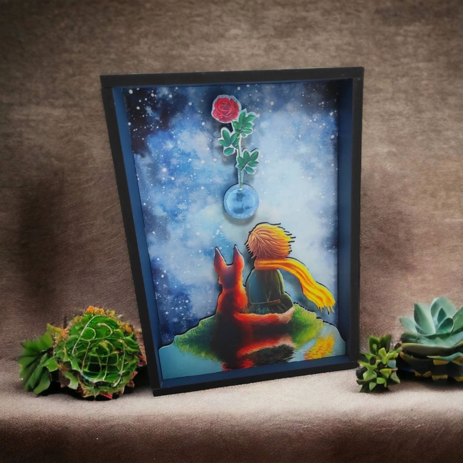 A framed artwork depicting a person and a dog sitting on a small island, looking at a starry night sky with a floating red rose in a glass vase above them. The scene includes vibrant colors and a dreamy atmosphere.
