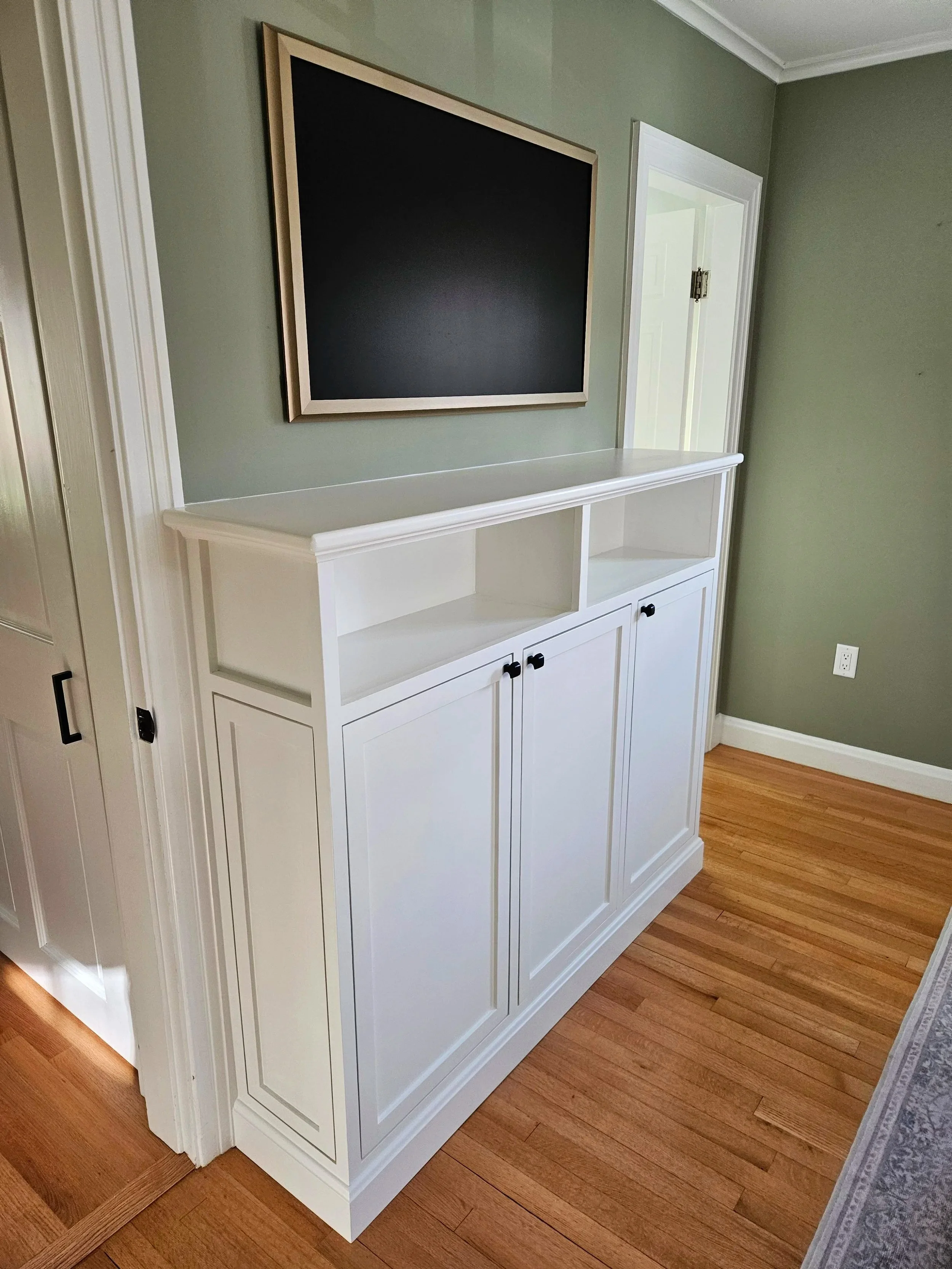 White built-in cabinet with closed doors and open shelves above, situated against a green wall with white trim, next to a door with white frame, and a hardwood floor.
