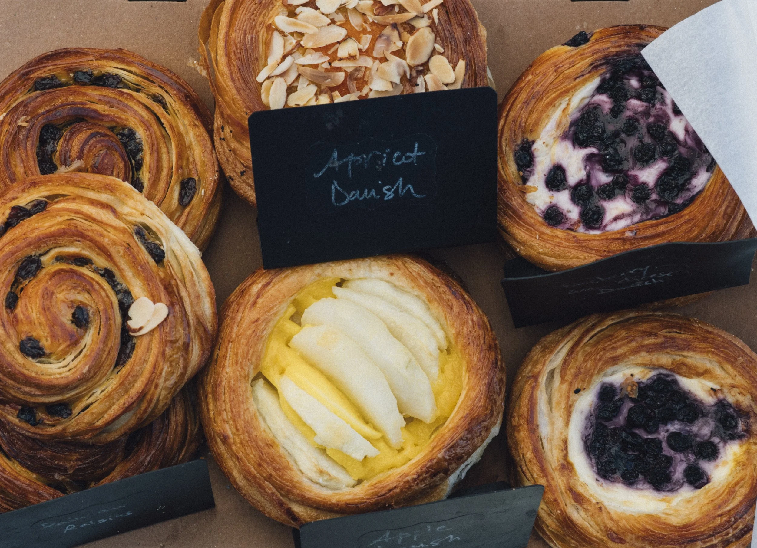 Assorted French pastries, including blueberry, almond, and lemon-flavored varieties, with small black chalkboard signs labeling them.