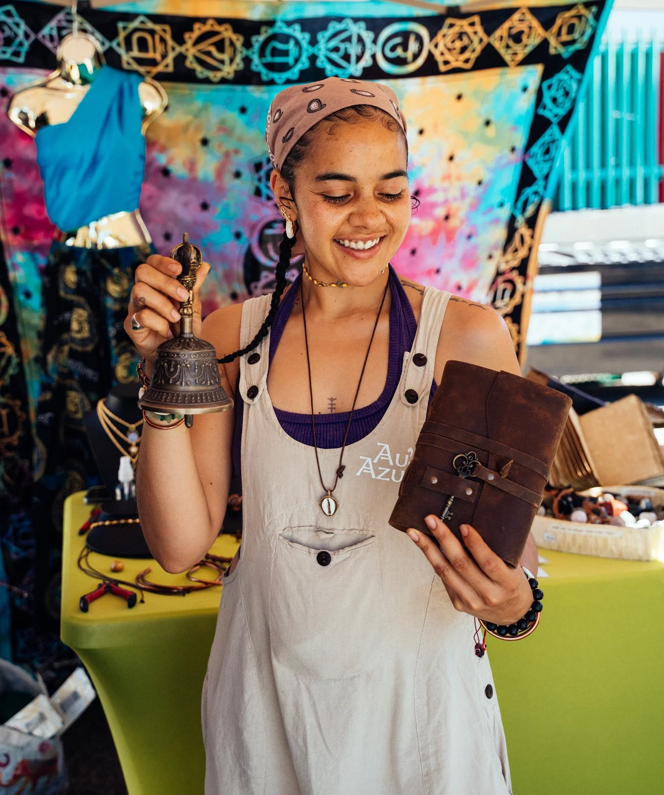 Young woman with braided hair and a head scarf smiling, holding a decorative bell in one hand and a leather journal with a key in the other, standing at a colorful arts and crafts booth