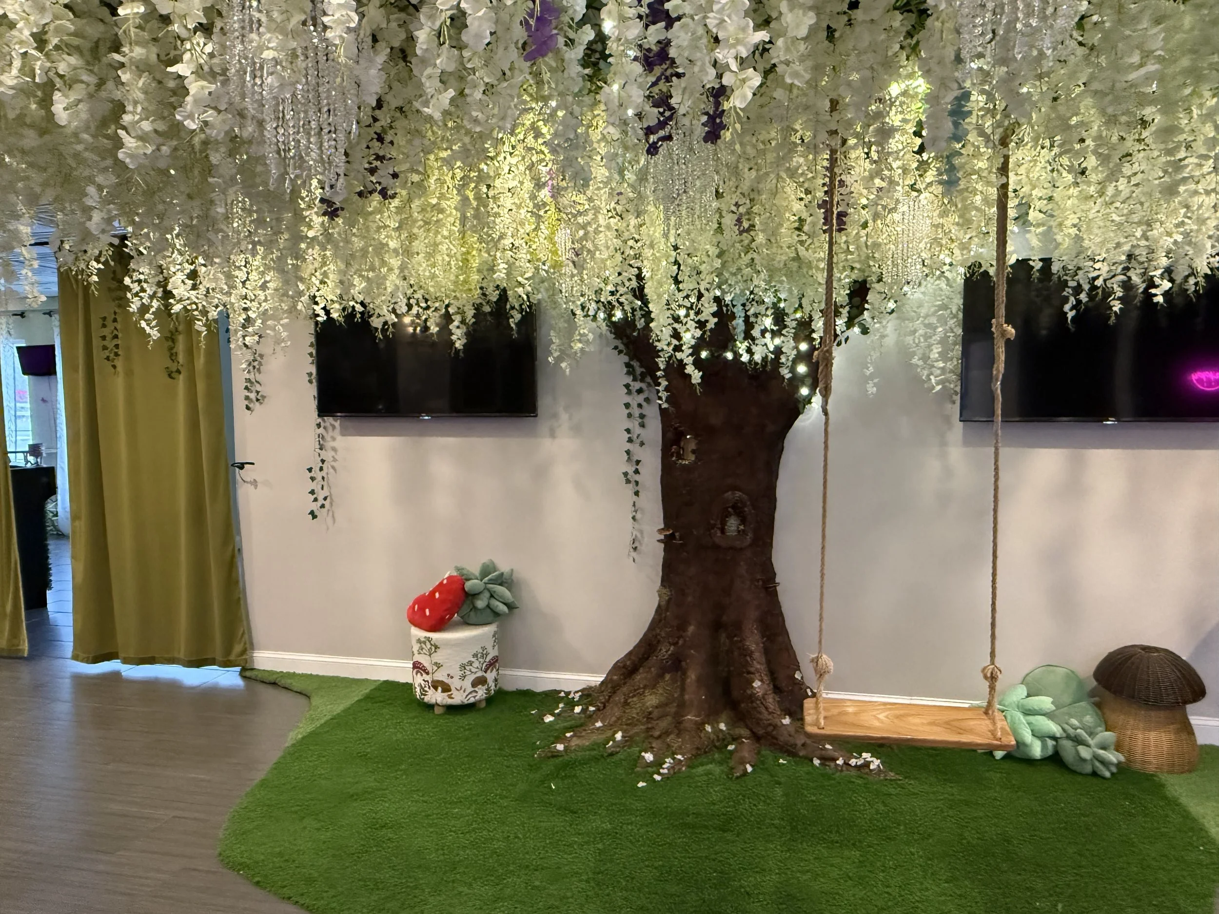 Indoor space decorated with a large artificial tree with hanging white and purple flowers, a swing hanging from branches, two flat-screen TVs on the wall, green curtains, a small mushroom-shaped wicker stool, a flower-patterned container with plush t