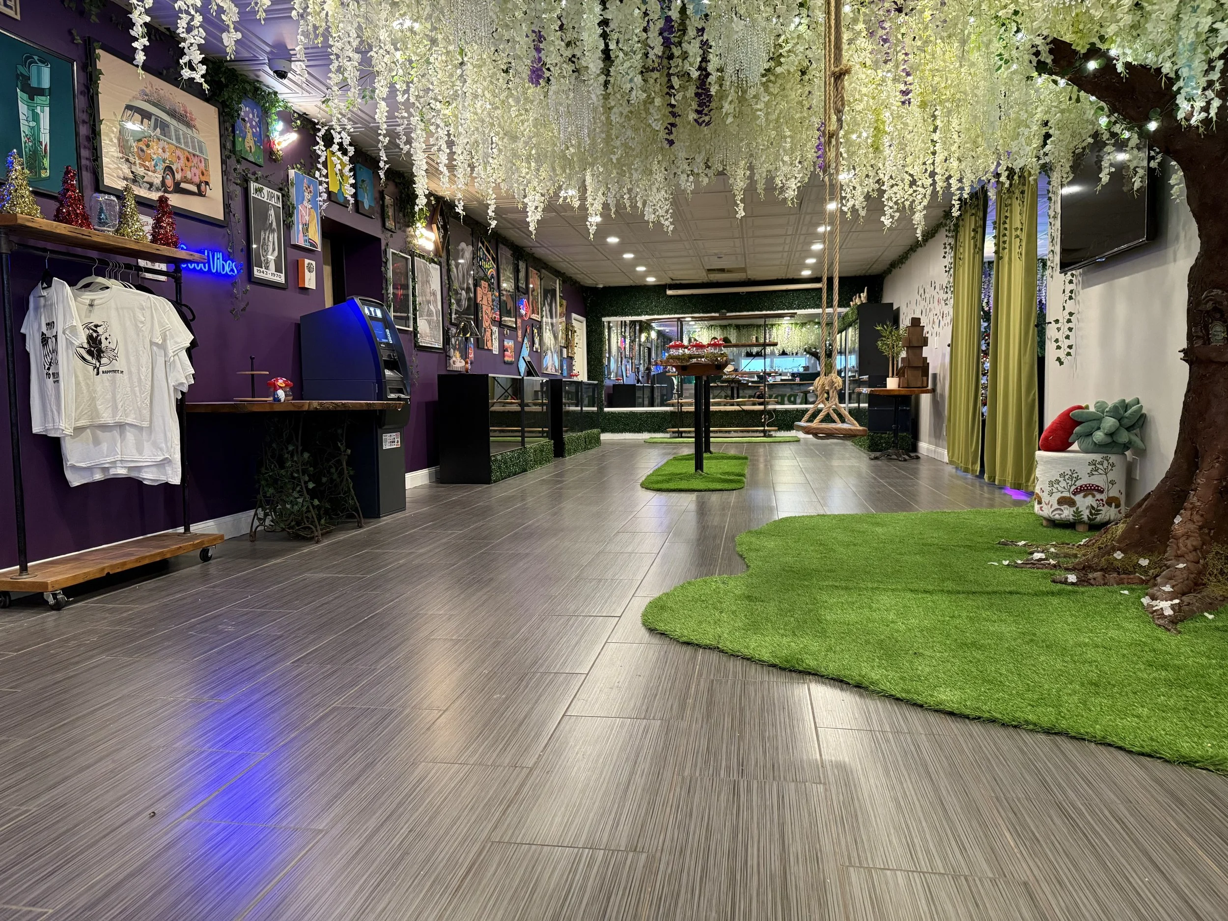 Indoor space decorated with hanging white and purple flowers, green artificial grass, trees, and colorful artwork on the walls, with a counter, clothes, and arcade machine on the left and a play area with swings and plants on the right.