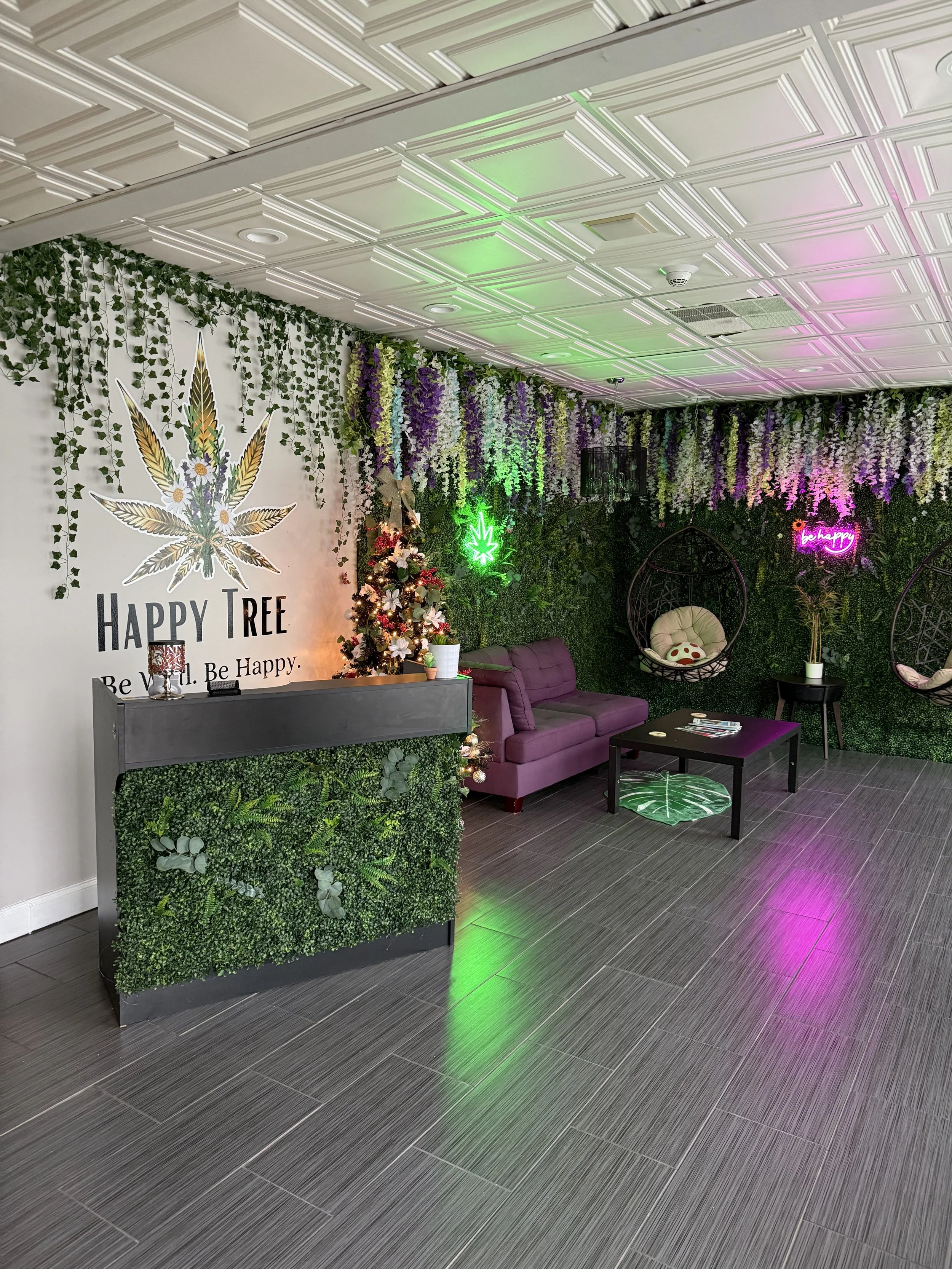A decorated indoor lounge area with a green and purple plant wall, neon signs, hanging chairs, a purple sofa, a small table, and a fake Christmas tree, creating a cozy, tropical, and modern ambiance.