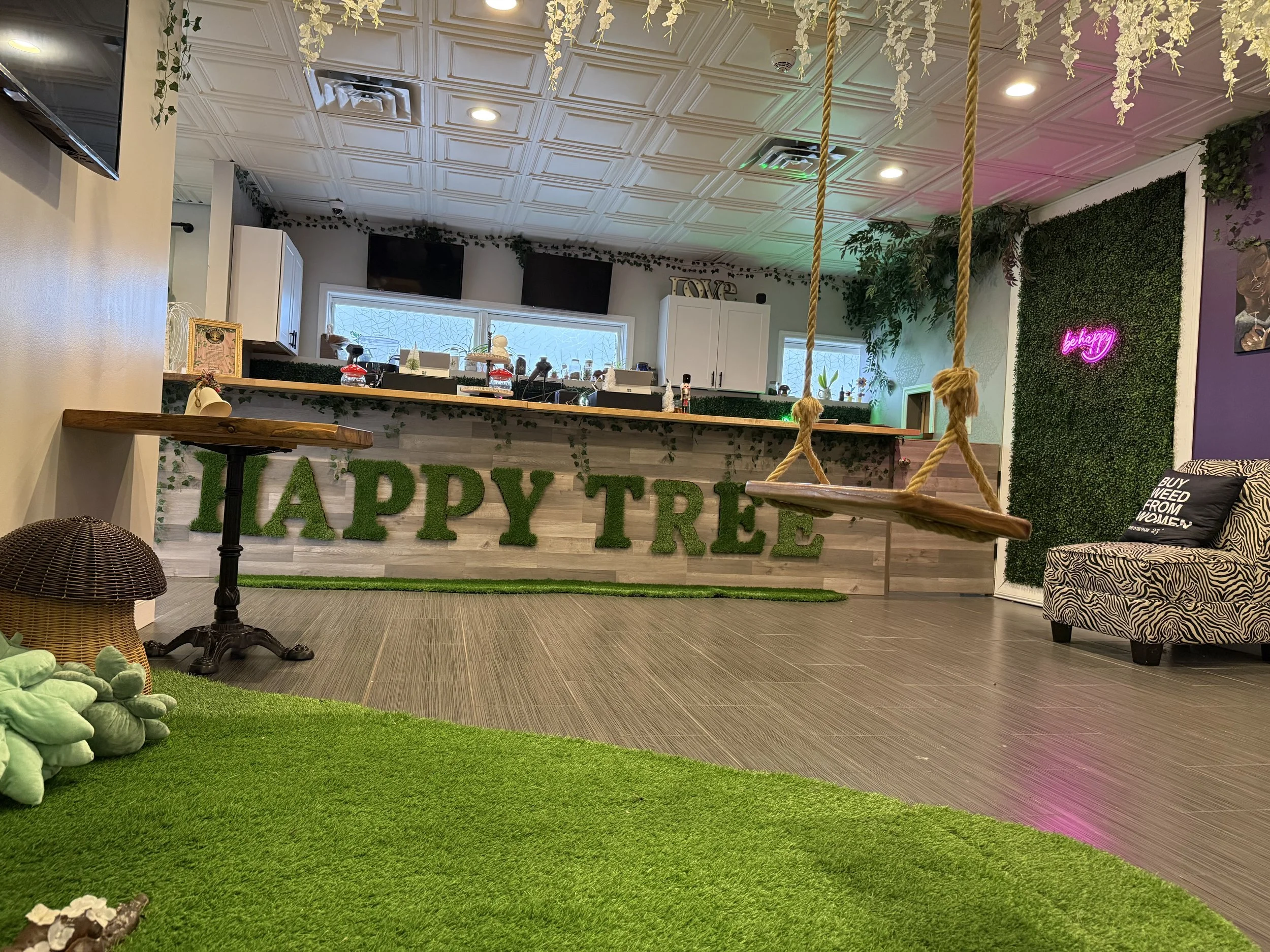 Indoor space with a green sign reading 'HAPPY TREE' on a wooden counter, two swings hanging from the ceiling, a zebra-print chair with a pillow that says 'BUY WEED FROM WOMEN,' and artificial grass and plants decorating the area.