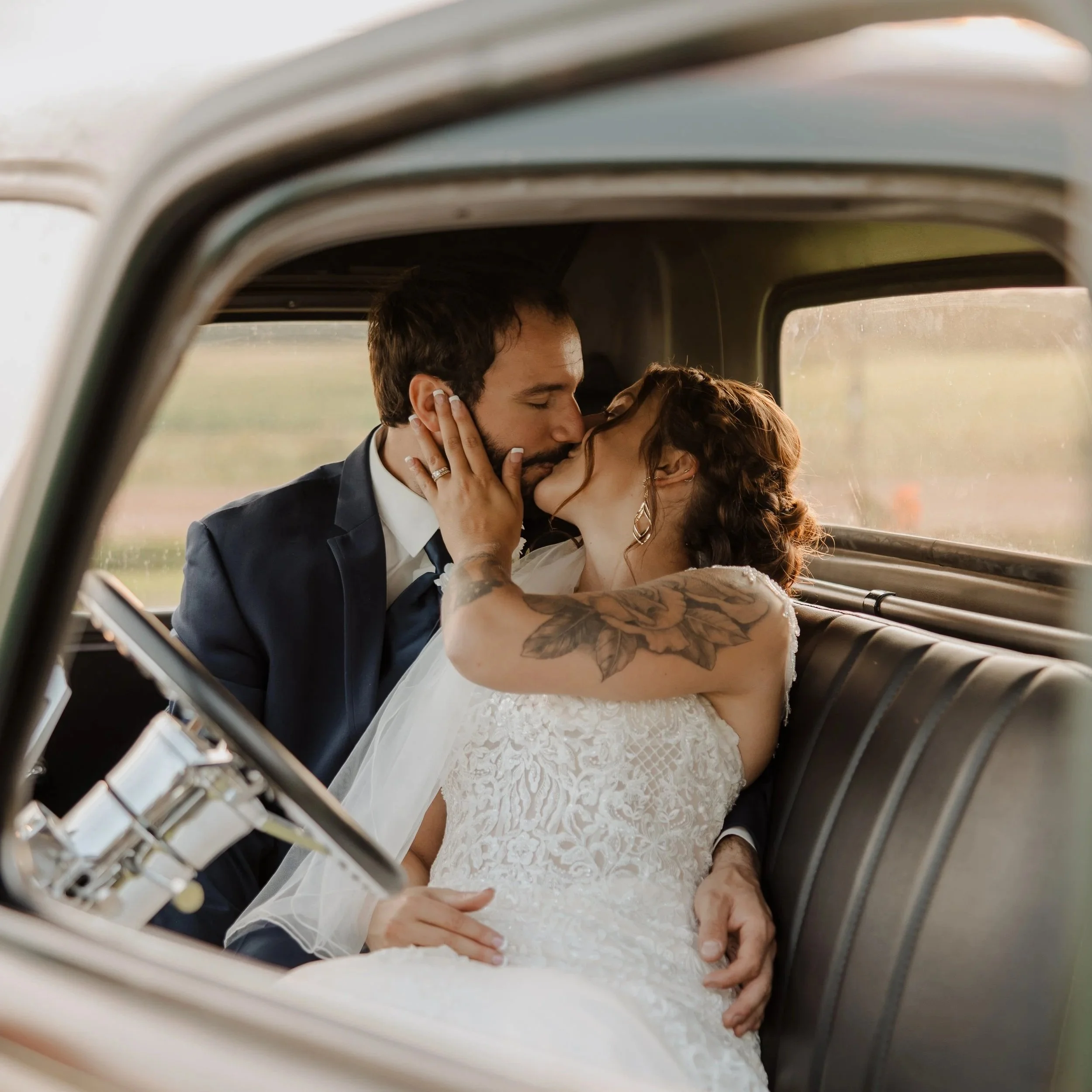 A bride and groom sharing a kiss inside a vehicle, visible through a small window frame.