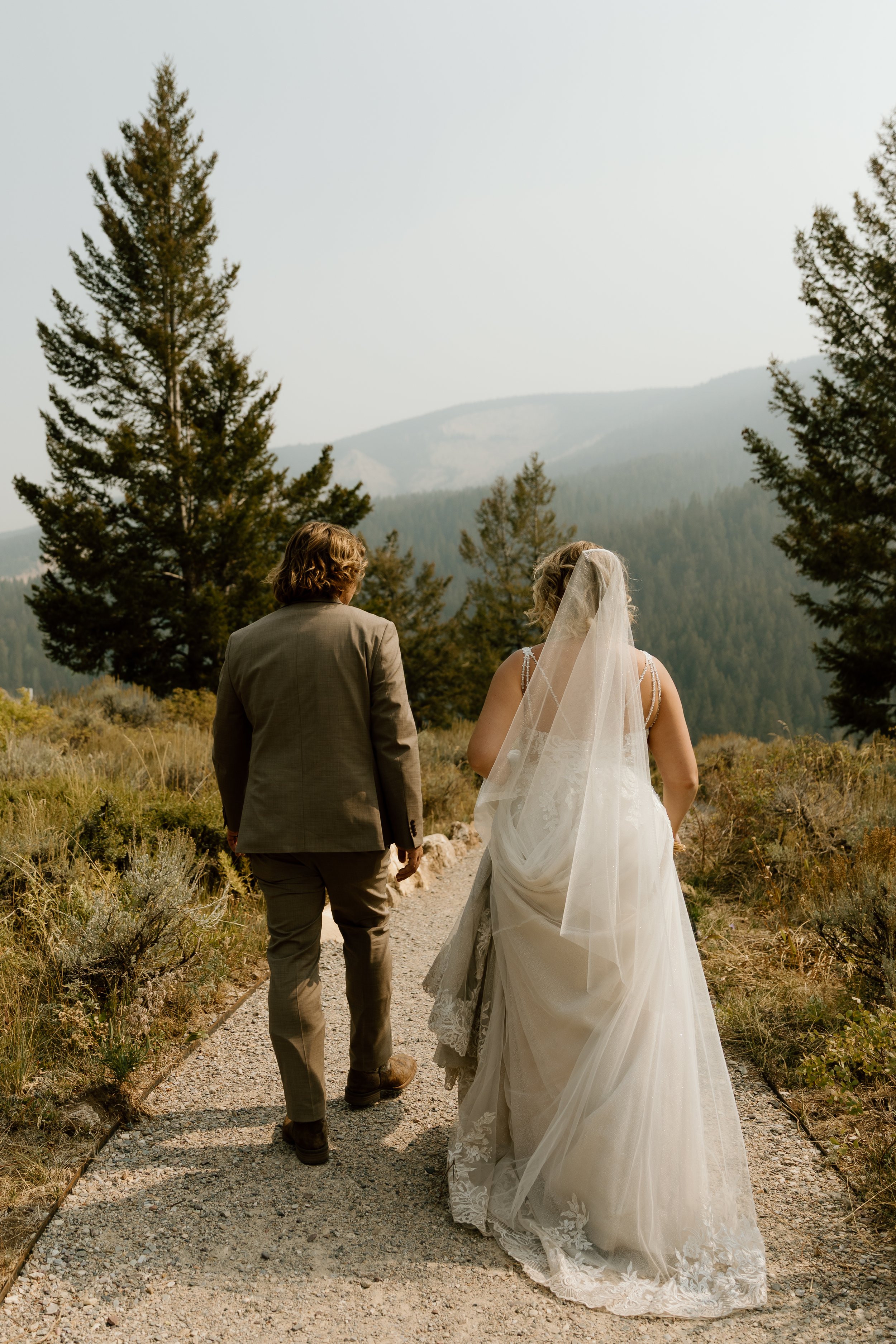 A bride and groom walking on a dirt path in a wilderness area with mountains and pine trees in the background, the bride wearing a white wedding gown and veil, the groom in a beige suit.