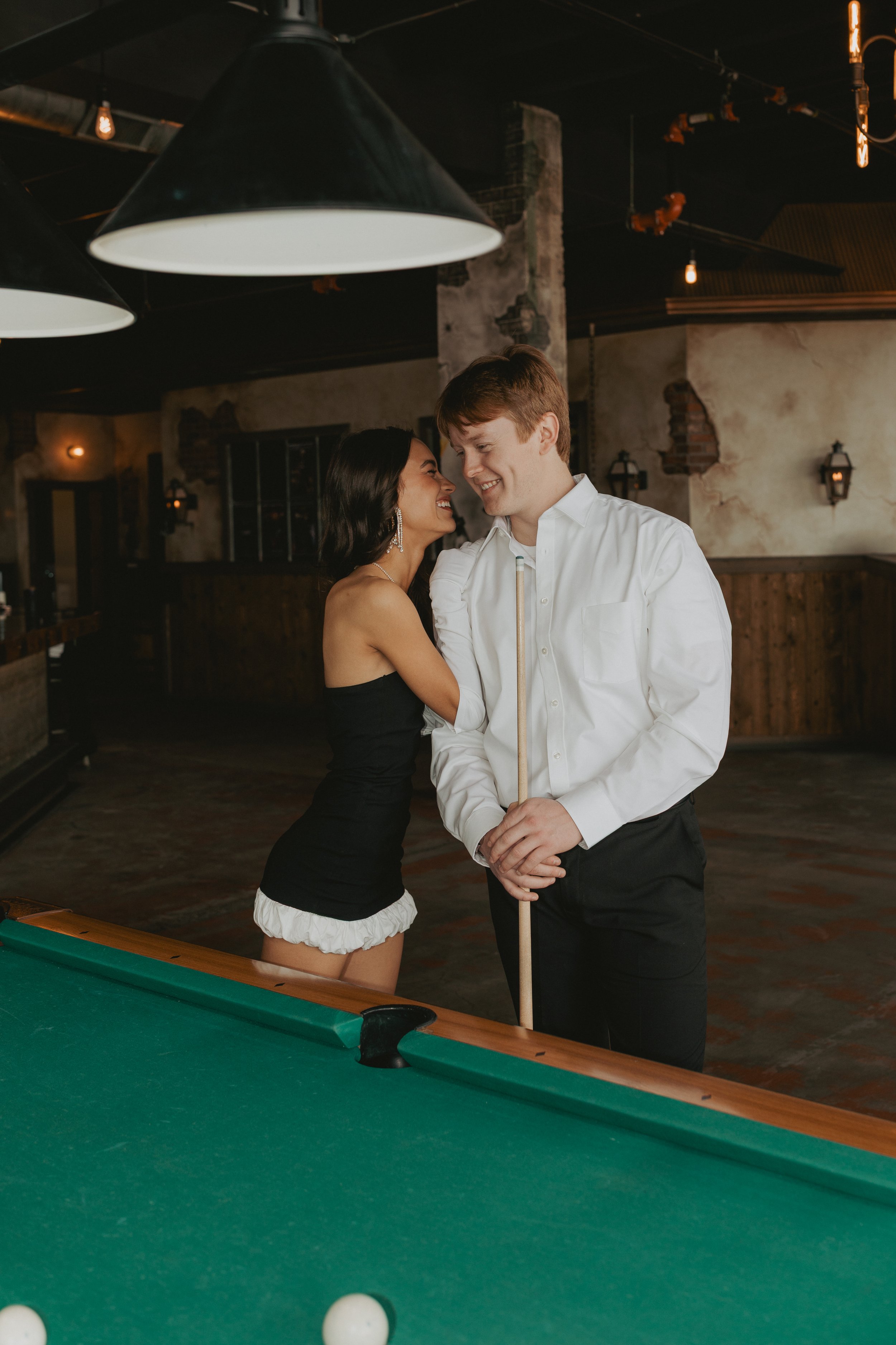 A woman and a man smiling at each other while standing by a pool table in a dimly lit bar, with the woman wearing a black top and white ruffled shorts, and the man in a white shirt holding a pool cue.
