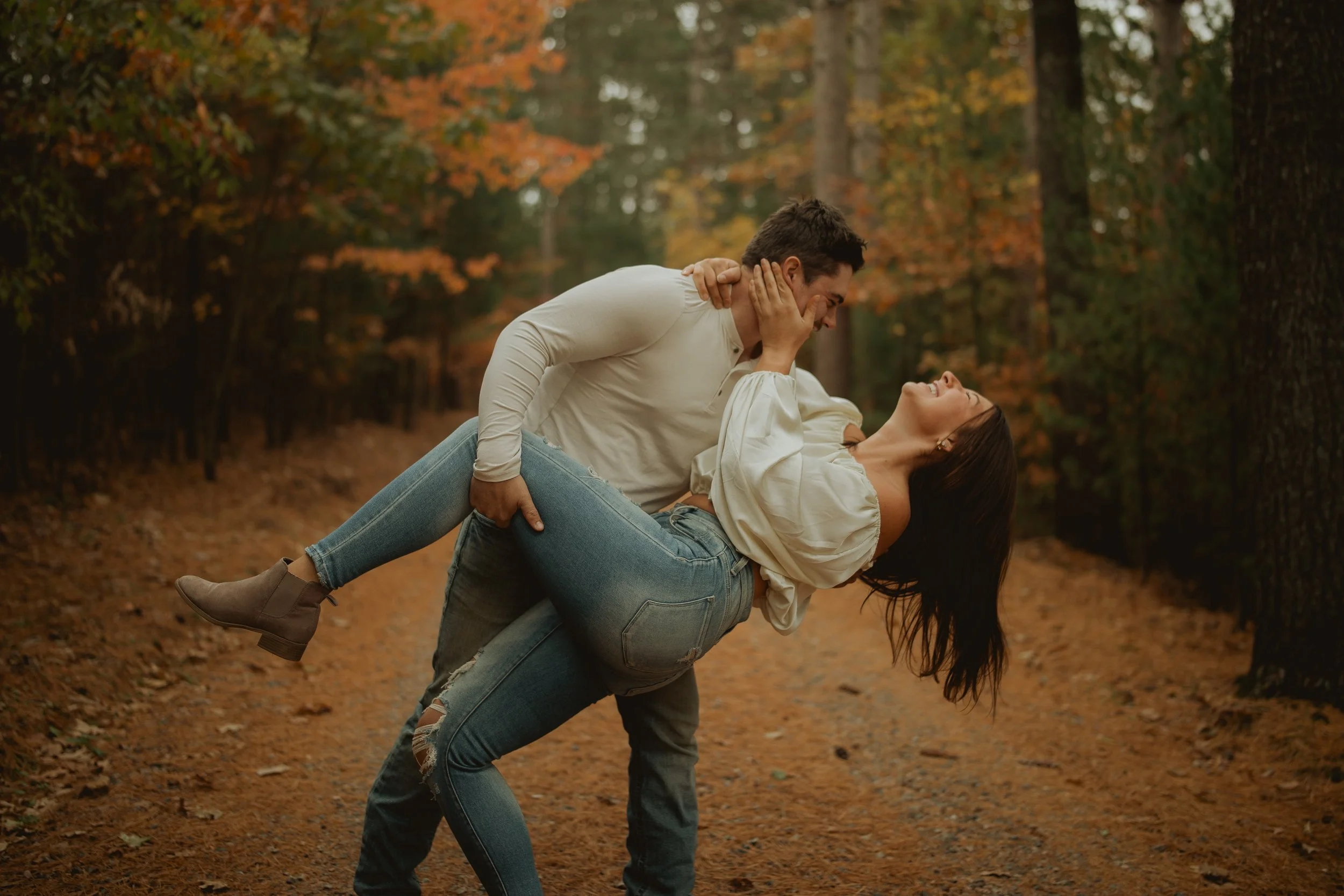 A man carrying a woman in a forest with autumn foliage, dressed casually, both smiling and enjoying each other's company.