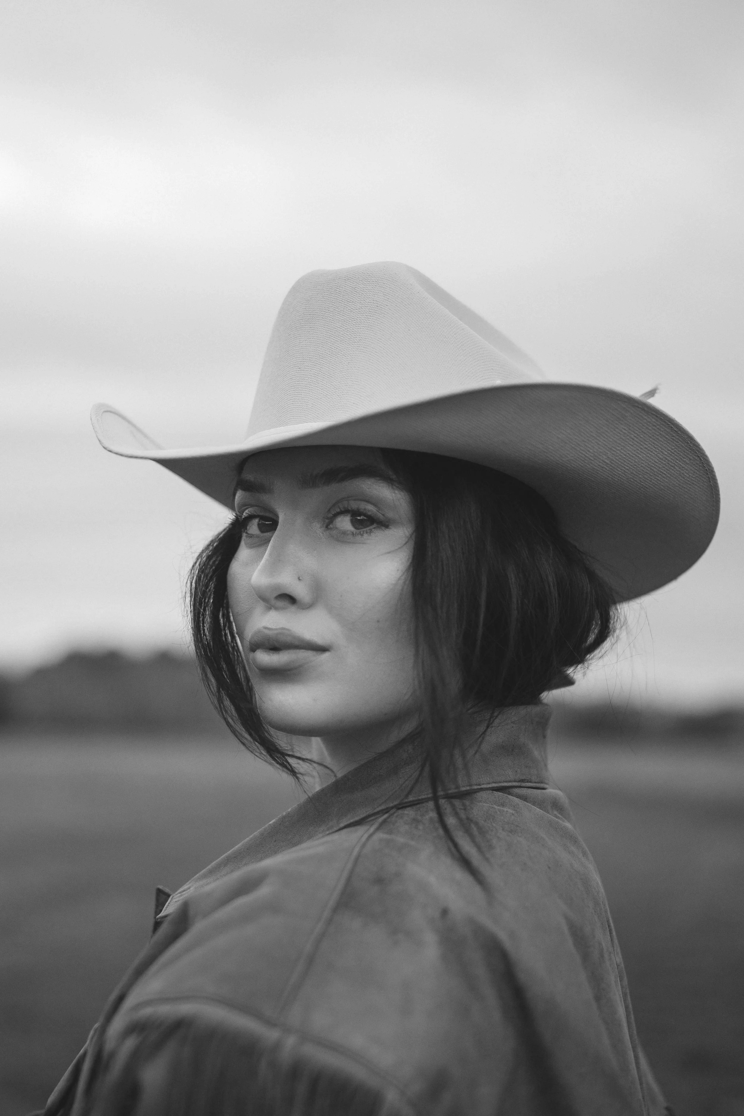 A woman with dark hair wearing a cowboy hat and a jacket, looking over her shoulder in an outdoor setting.
