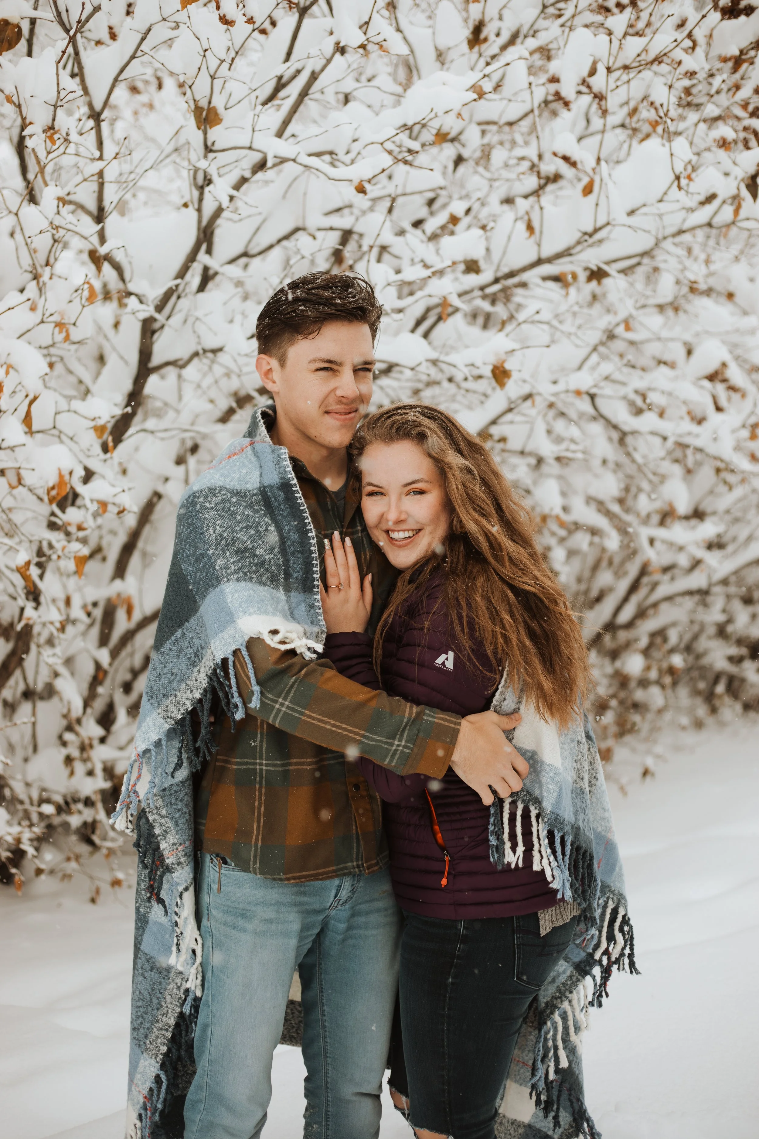 A young man and woman standing close together snowy winter landscape, with snow-covered trees behind them. They are smiling and hugging, wearing cozy winter clothing and a blanket draped over their shoulders.