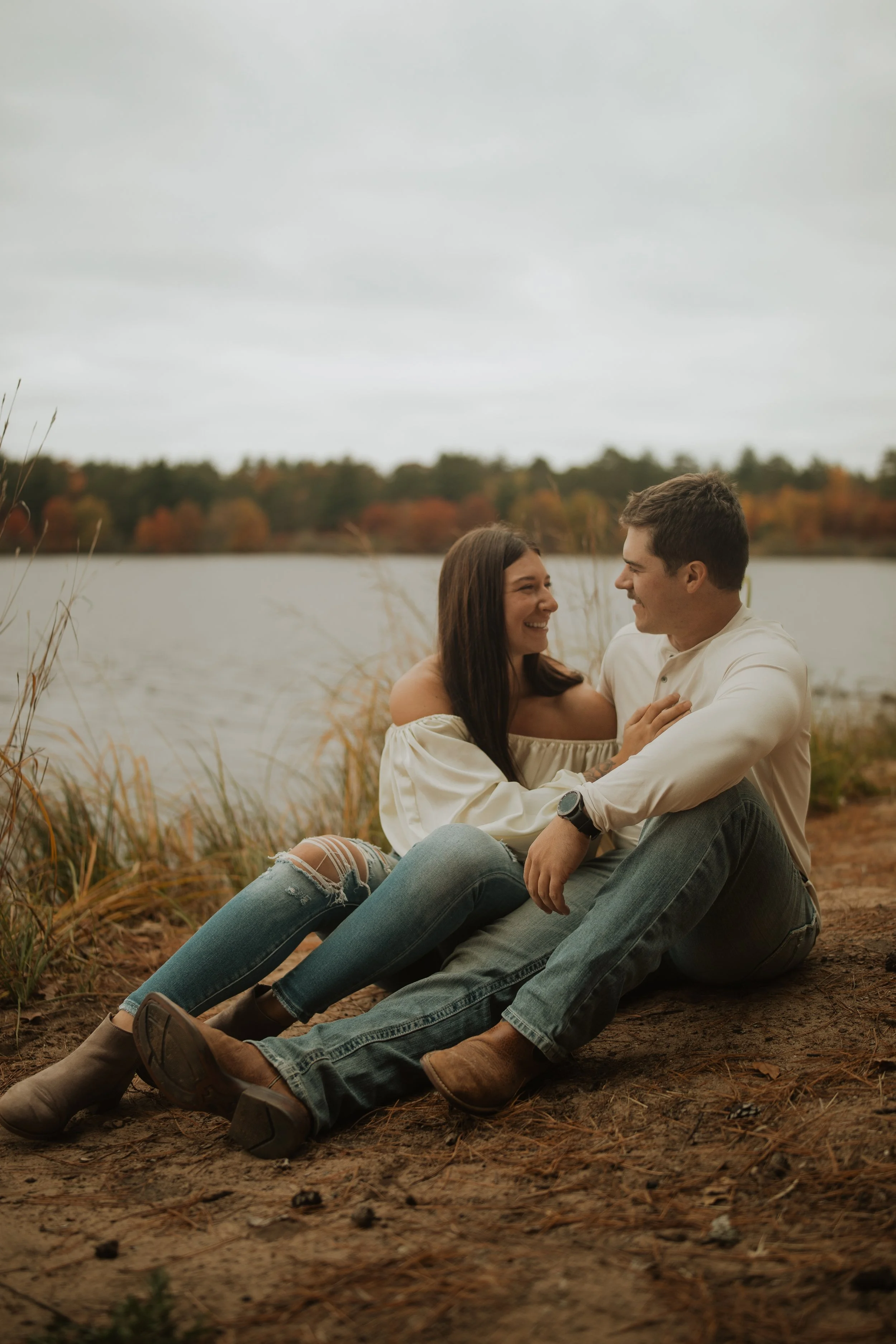 A couple sitting on the ground near a lake, smiling at each other during an overcast day, with a background of fall-colored trees.