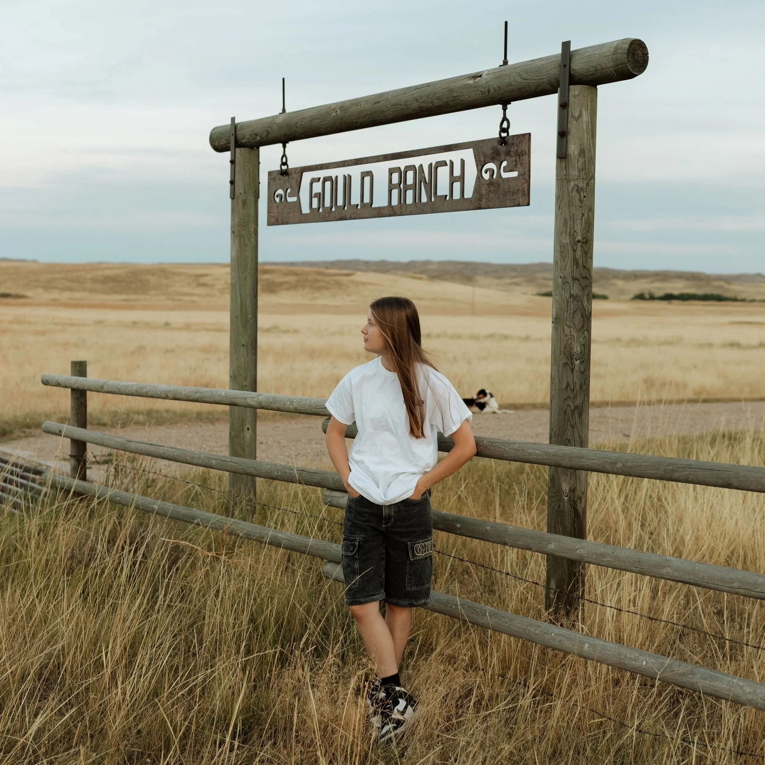 A young woman with long brown hair wearing a white t-shirt and black shorts standing by a wooden fence in a rural area. Behind her, a sign reads 'GOLD BUNCH' and is attached to a wooden post, with a landscape of open fields and rolling hills under a cloudy sky.
