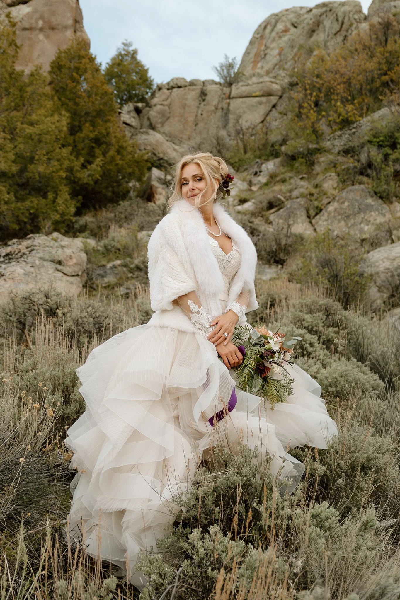 A woman in a white wedding dress with lace details and a layered tulle skirt, wearing a white fur stole and a pearl necklace, holding a bouquet of flowers, standing outdoors in a rocky, desert-like landscape with bushes and trees, and large rock formations in the background.