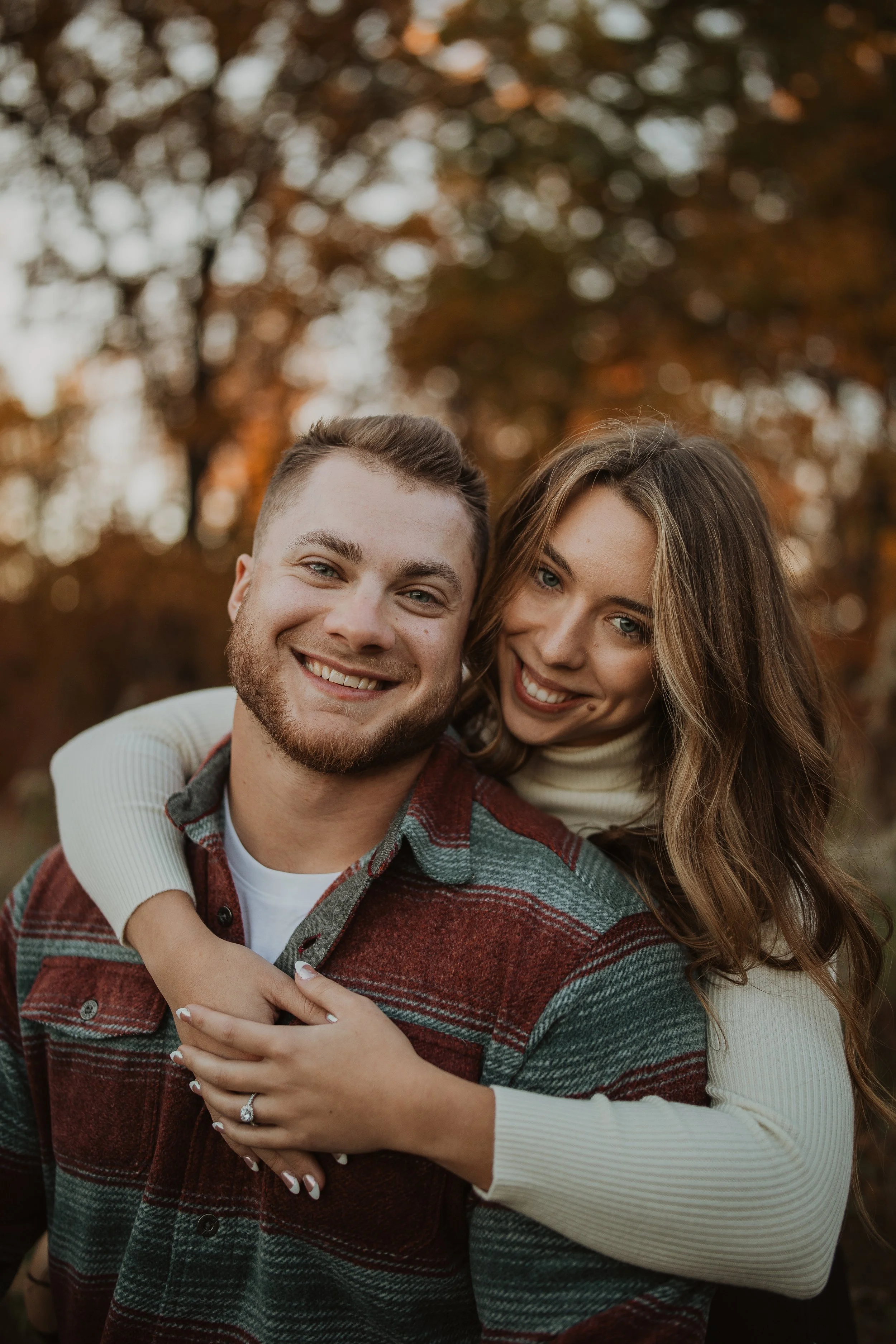 Close-up of a couple smiling outdoors during fall, with autumn trees in the background. The woman has long, wavy hair and is wearing a beige turtleneck, and the man has short hair and a beard, wearing a plaid shirt with red, gray, and green tones.