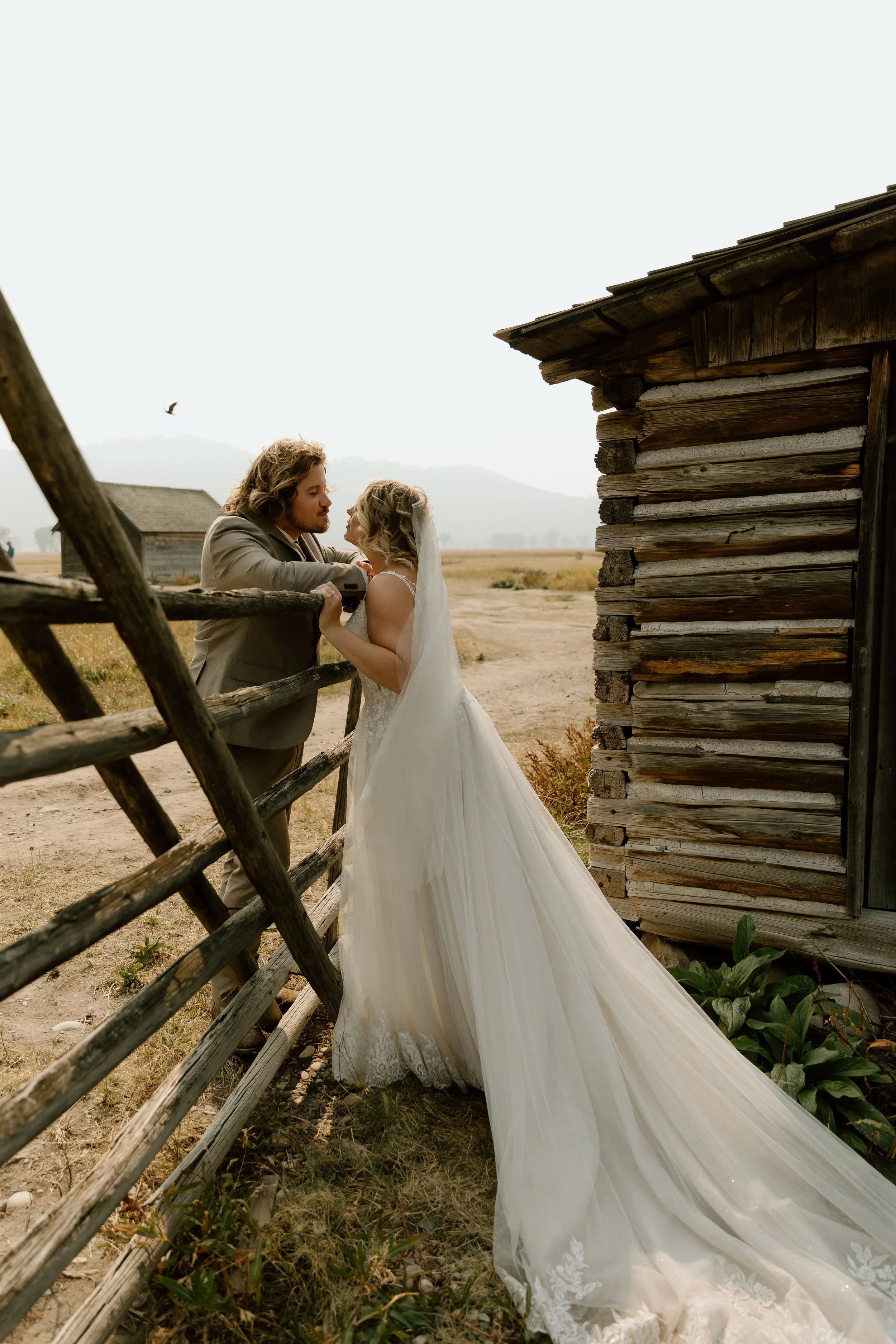 A bride and groom sharing a moment in a rural setting with an old wooden fence and a weathered wooden building nearby.