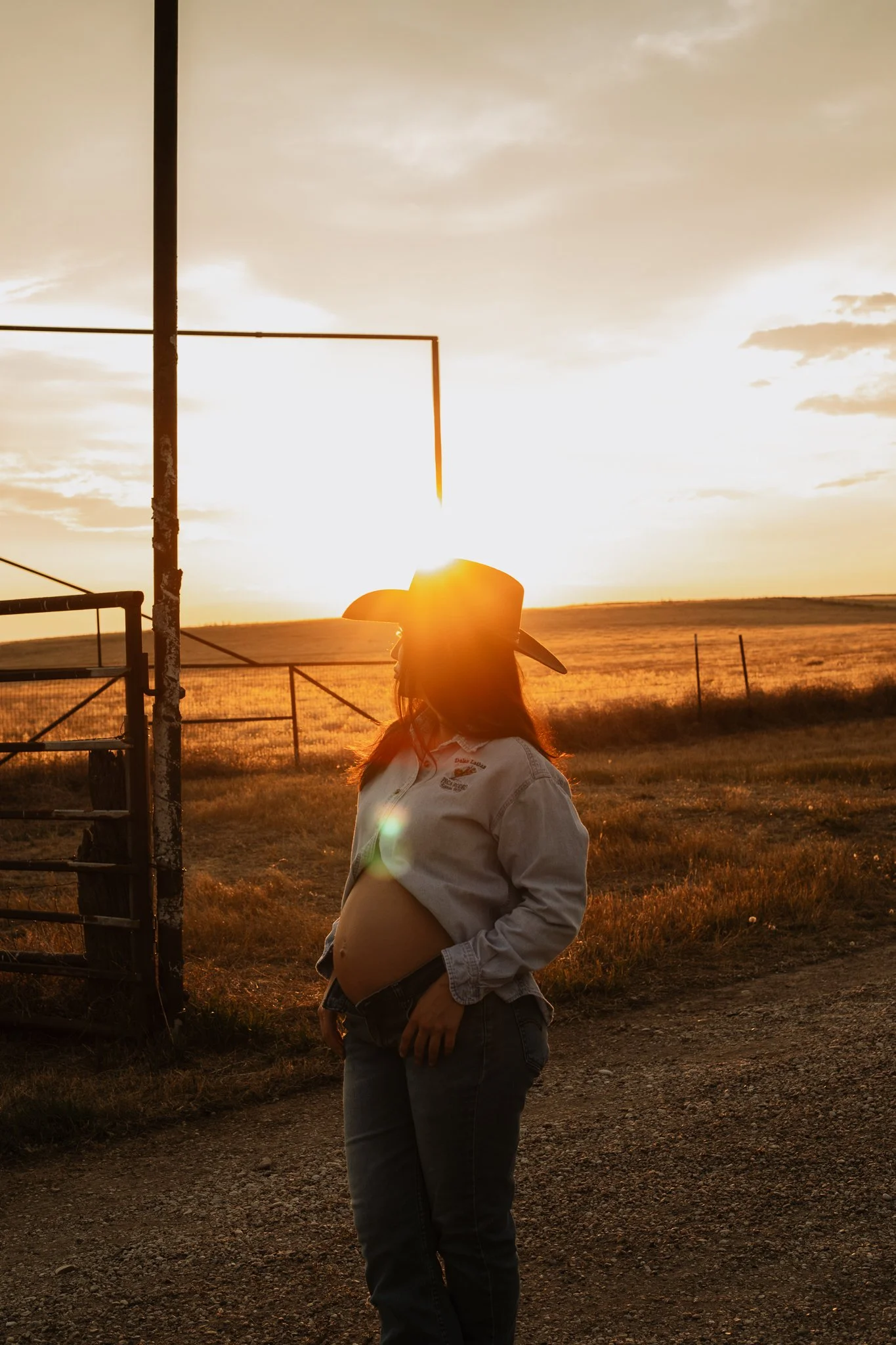 A pregnant woman wearing a cowboy hat and denim jacket, holding her belly, standing outdoors at sunset near a rural fence.