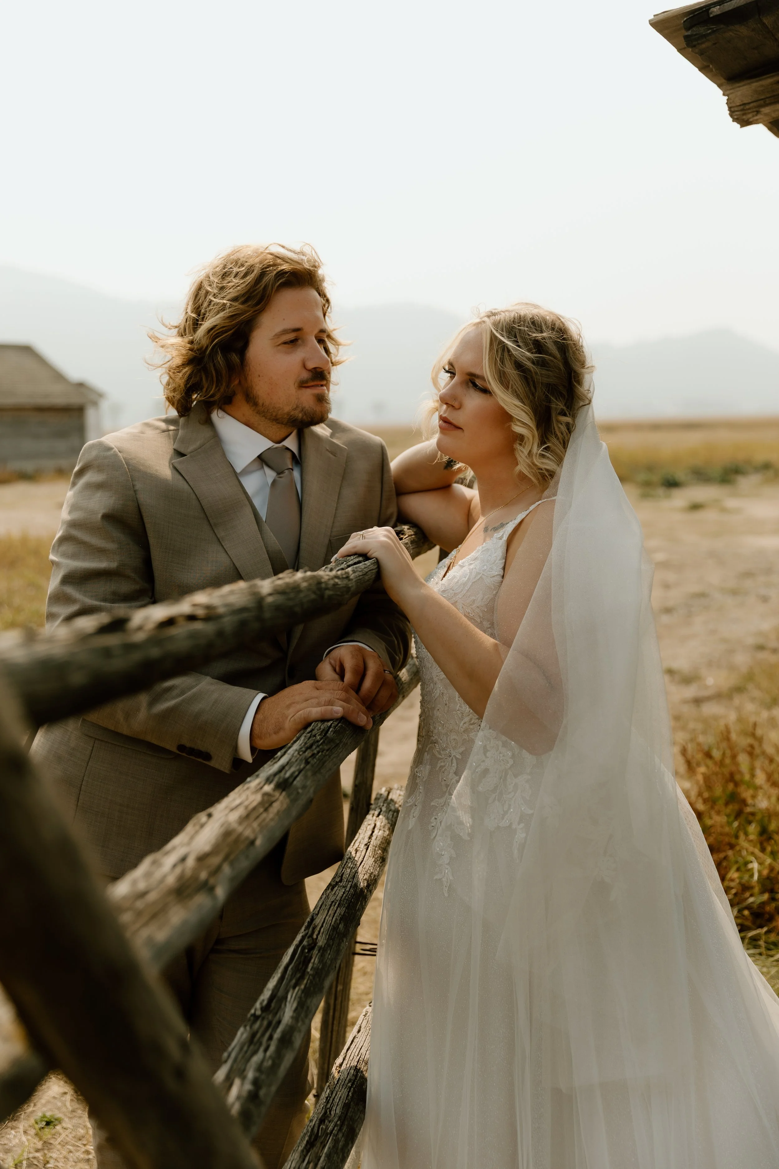 A bride and groom in wedding attire leaning on a rustic wooden fence outdoors with a rural landscape and the Grand Tetons in the background.