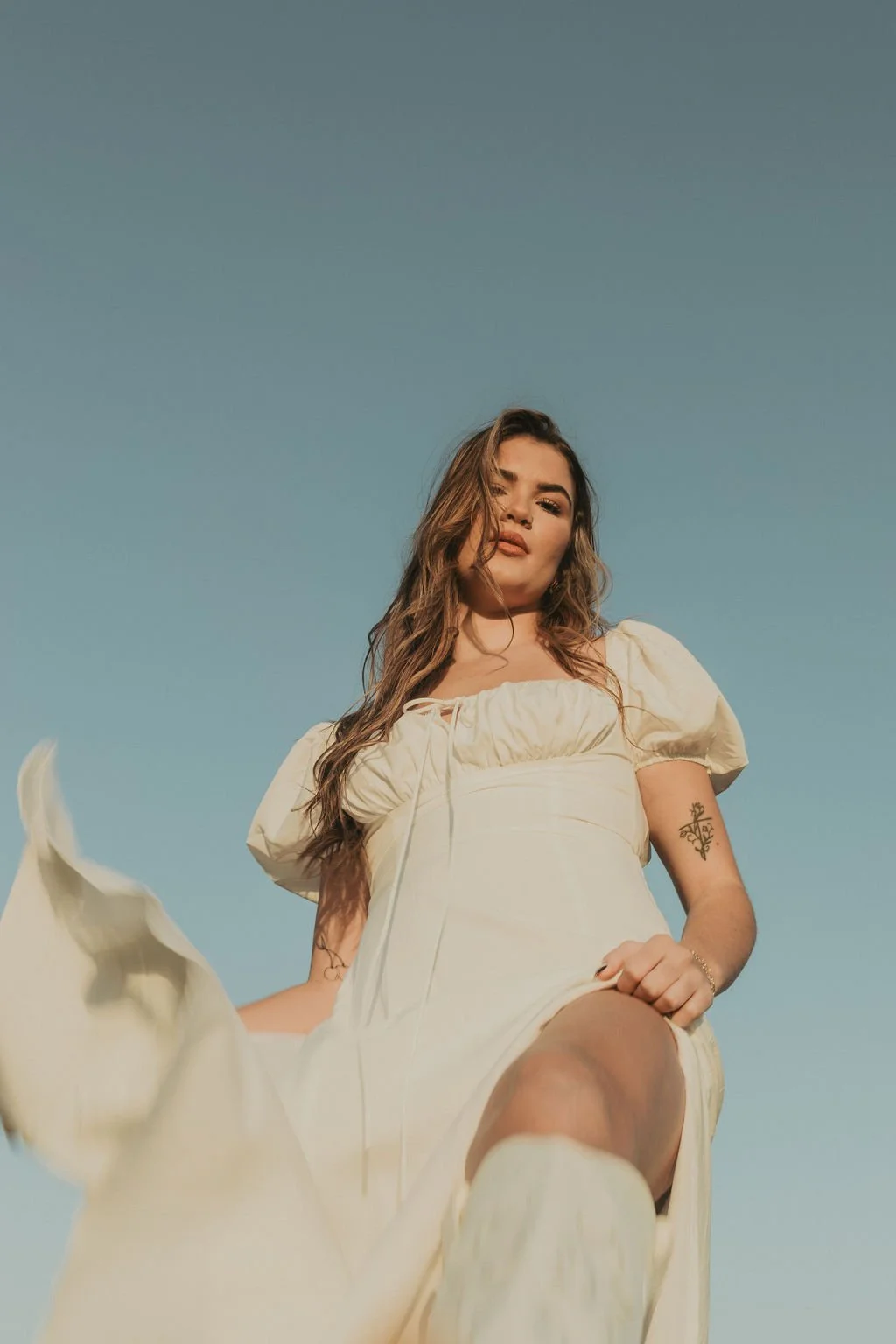 Young woman in cream-colored dress with puffed sleeves, standing against a clear blue sky, looking confidently at the camera.