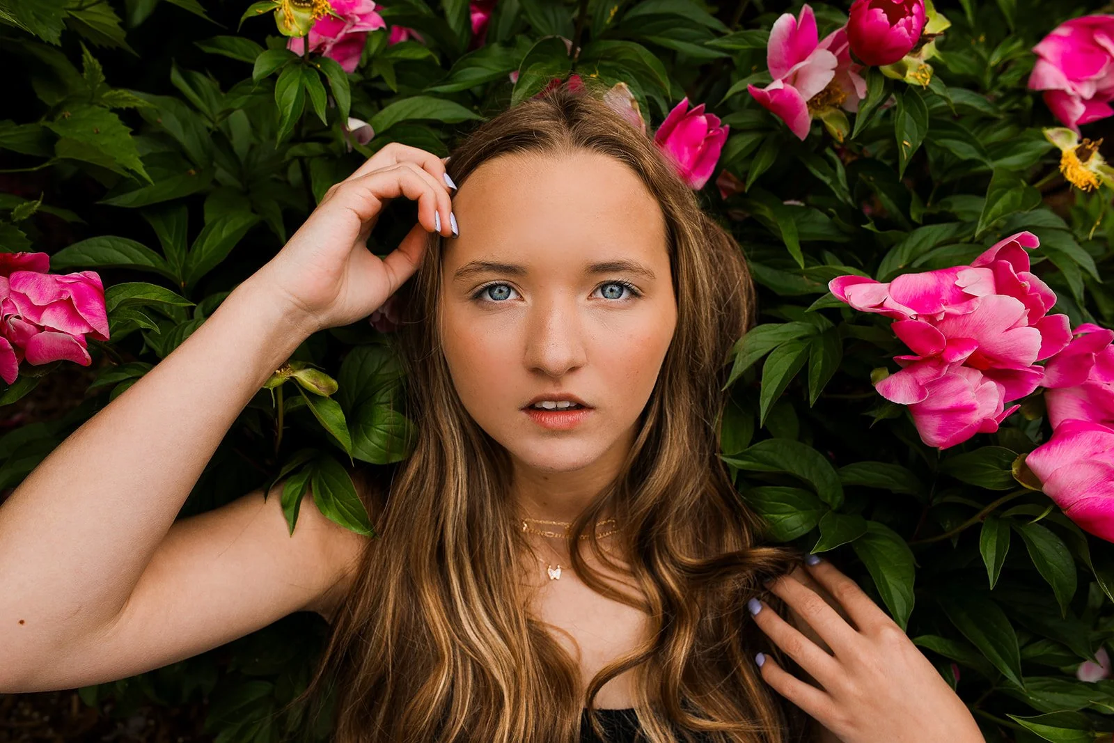 A young woman with long, wavy brown hair and striking blue eyes lying among pink and magenta peonies with green leaves, touching her hair and face.
