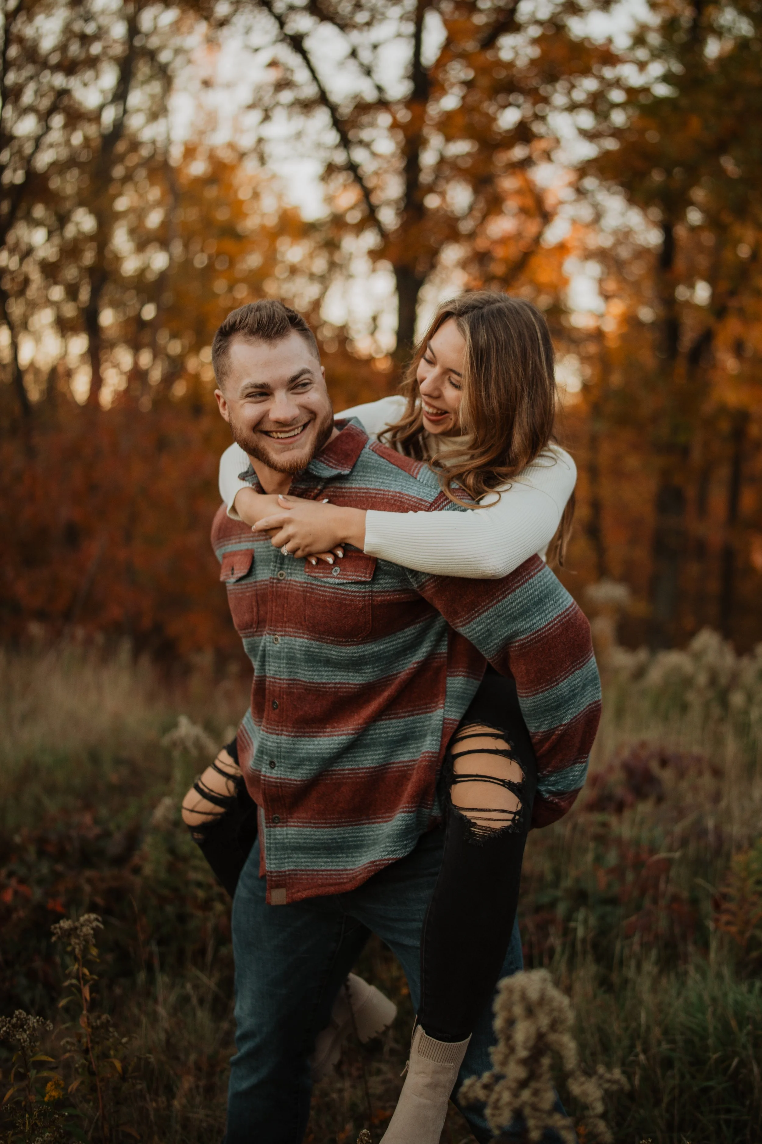 A happy couple in fall outdoor setting, the man giving the woman a piggyback ride, both smiling, surrounded by orange and brown autumn leaves.