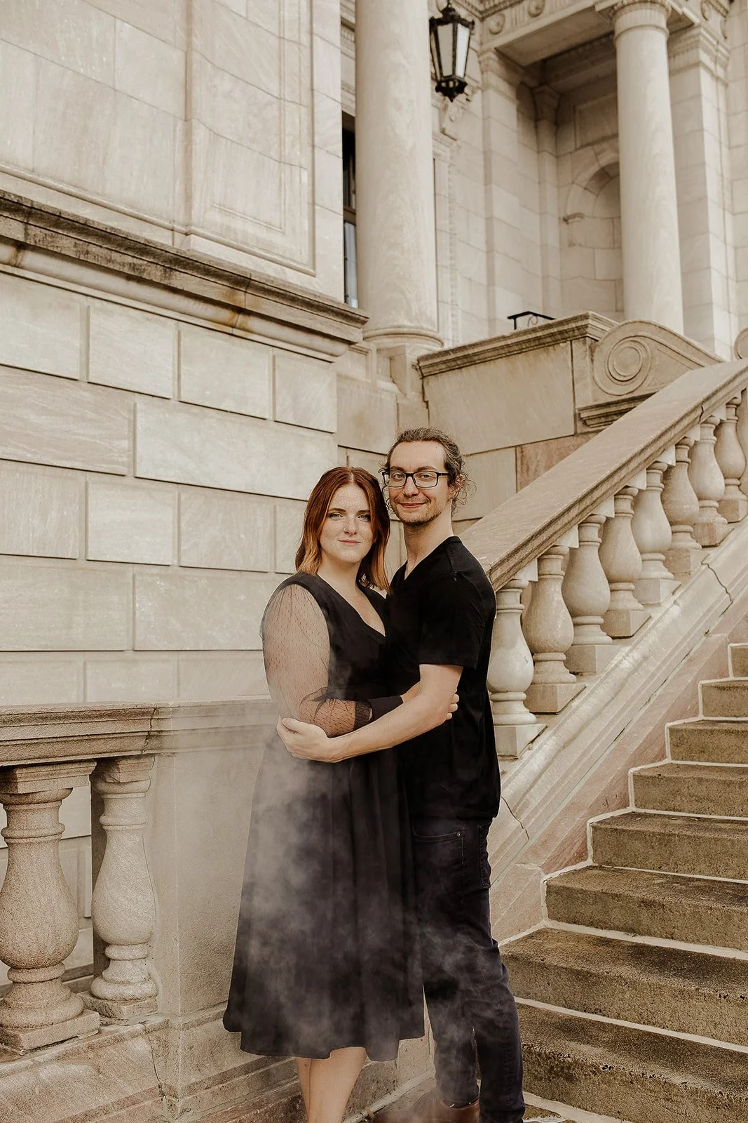 A couple stands on a stone staircase in front of a historic building with large columns and intricate architectural details. The woman has red hair and is wearing a black dress, and the man has glasses, long hair, and is dressed in black. They are embracing each other and smiling.