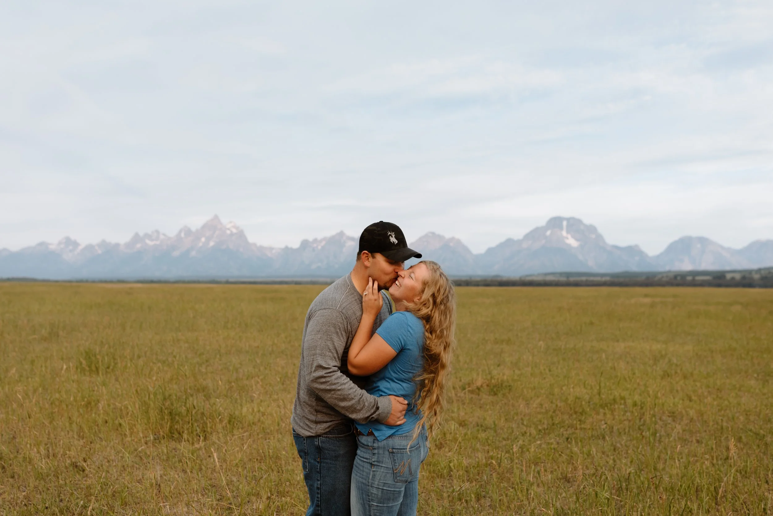 A couple standing in a grassy field holding each other and sharing a kiss, with the Grand Tetons in the background under a partly cloudy sky.