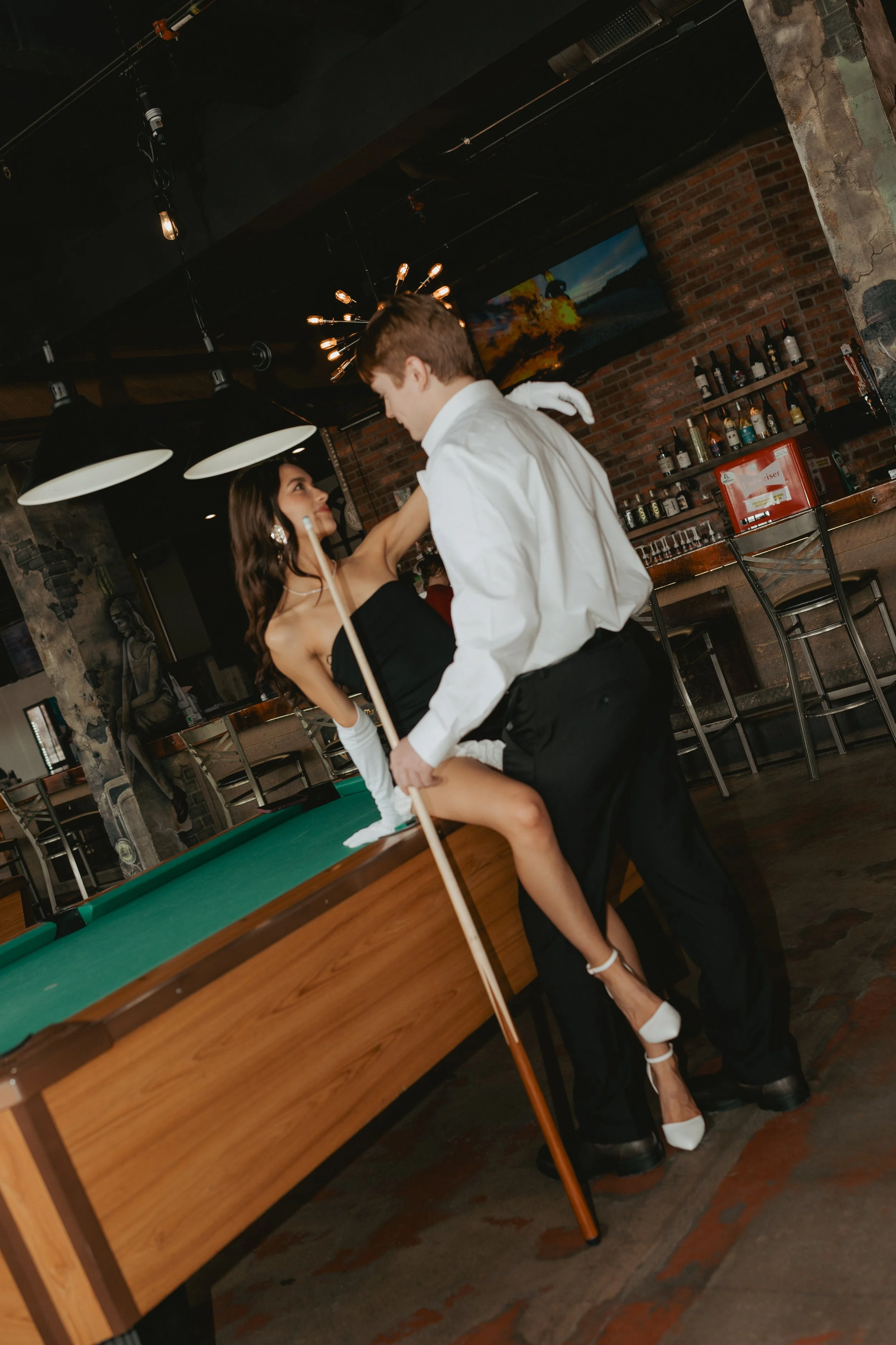A couple dancing in a bar, with the woman sitting on a pool table and the man standing next to her. The woman is wearing a black strapless dress and white heels, while the man is dressed in a white shirt and black pants. The bar has exposed brick walls, bottles on shelves, and a large painting of a fiery explosion in the background.