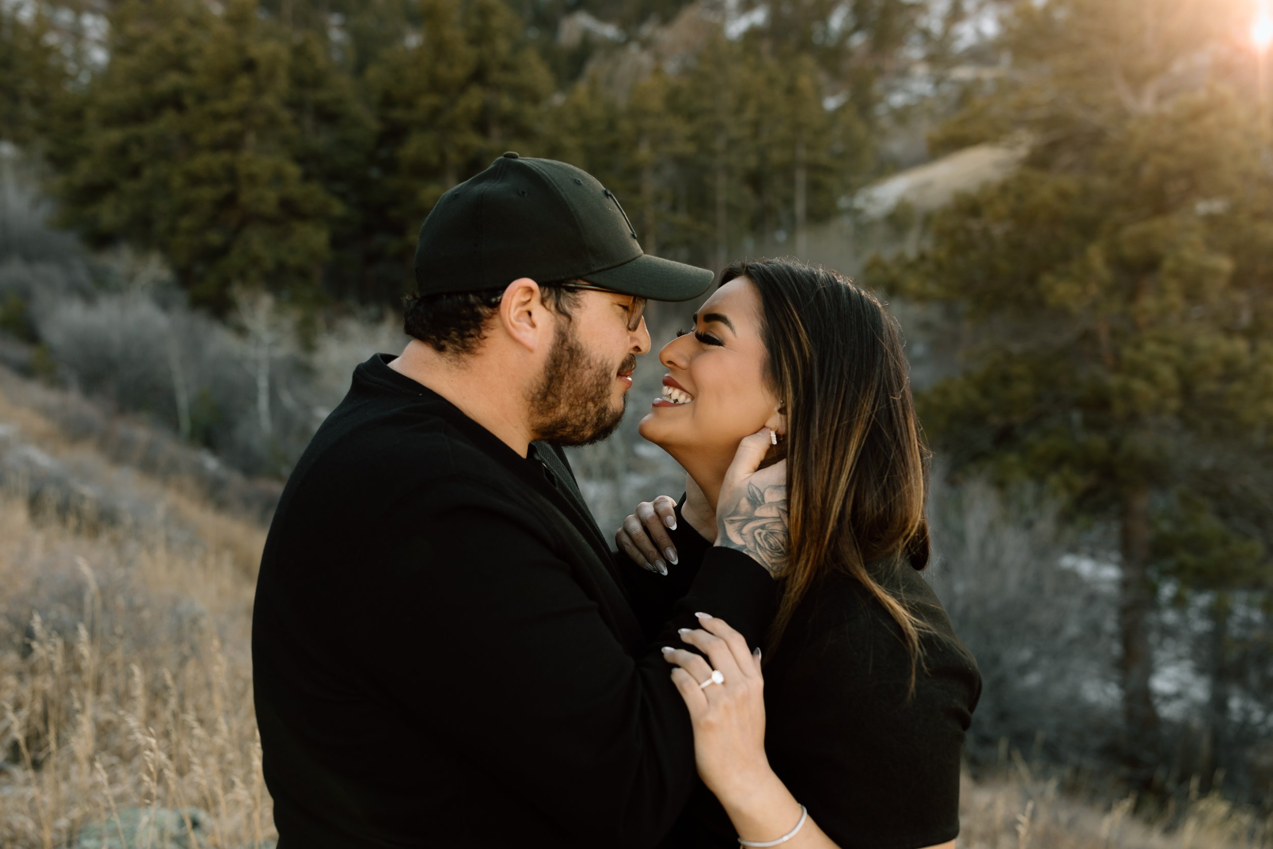 A couple faces each other closely outdoors with trees and sunlight in the background, smiling and embracing.