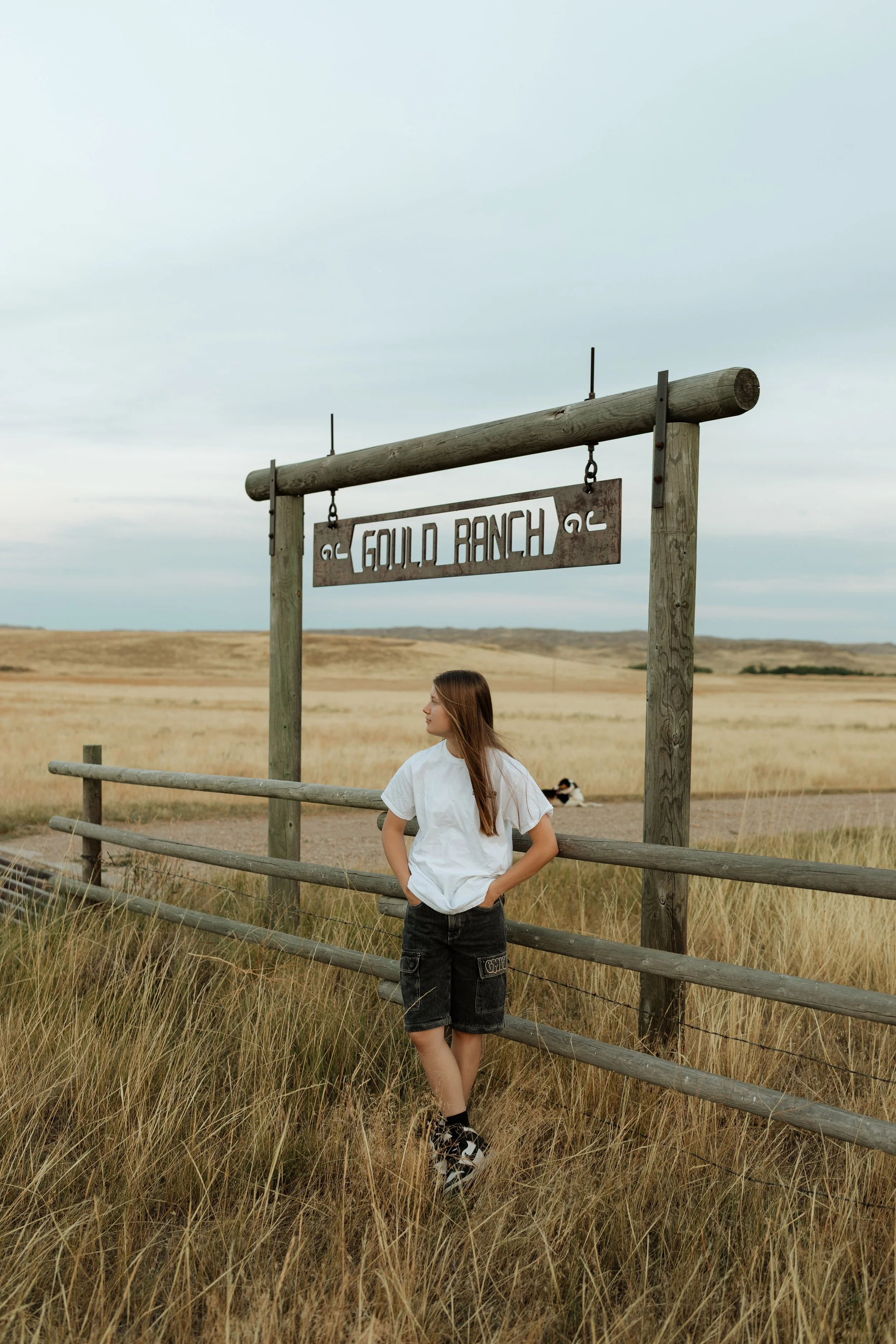 A young woman with long brown hair wearing a white t-shirt and black shorts, standing next to a wooden fence in a rural landscape with tall grass and rolling hills, under a cloudy sky, near a wooden sign that reads 'Gould Ranch'.