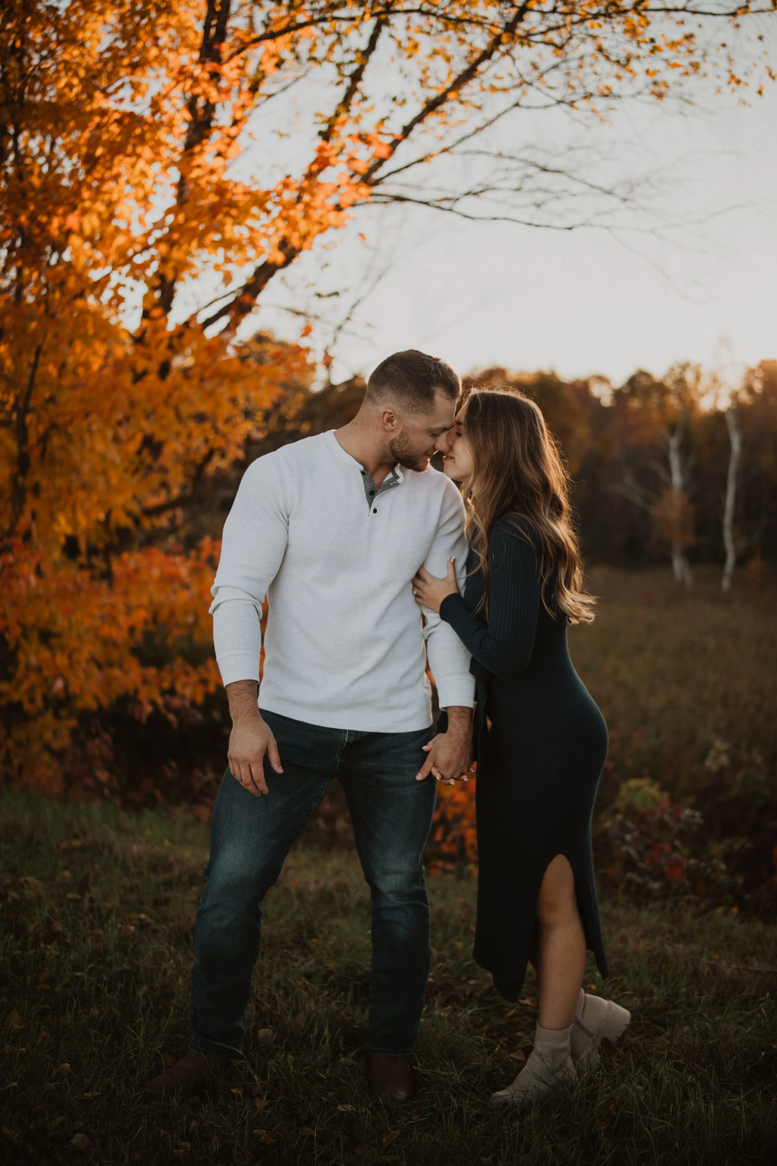 A couple holding hands and leaning close to each other outdoors during sunset, surrounded by fall foliage with orange and yellow leaves.
