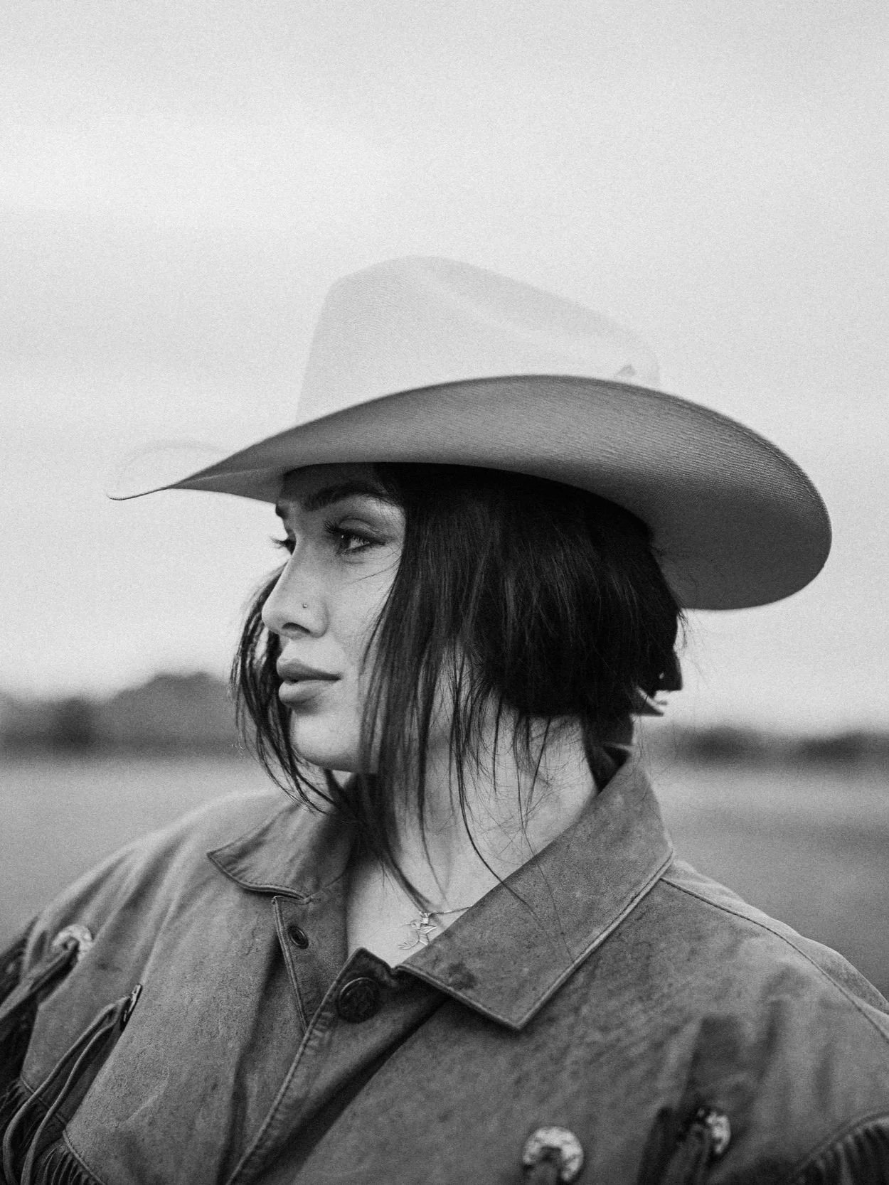 A black and white photo of a woman wearing a cowboy hat and a jacket, looking to the left with a blurred background.