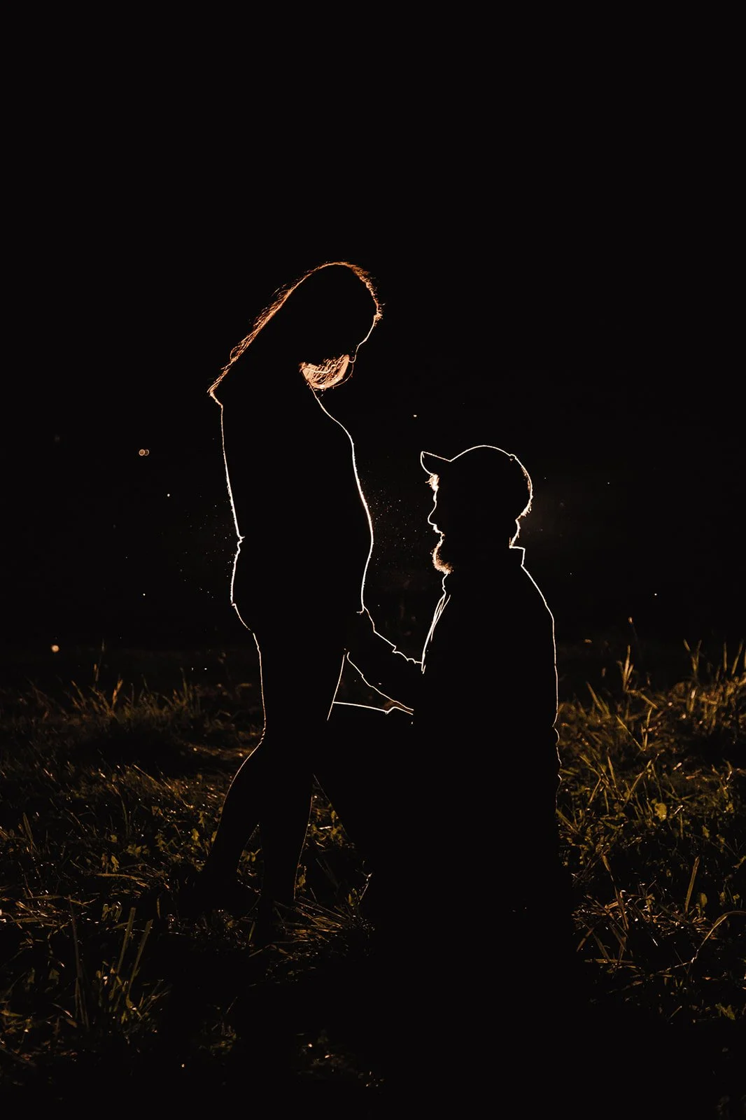 Silhouette of a couple at night, with the man kneeling and holding the woman's pregnant stomach, both standing on grass, illuminated by a backlight.