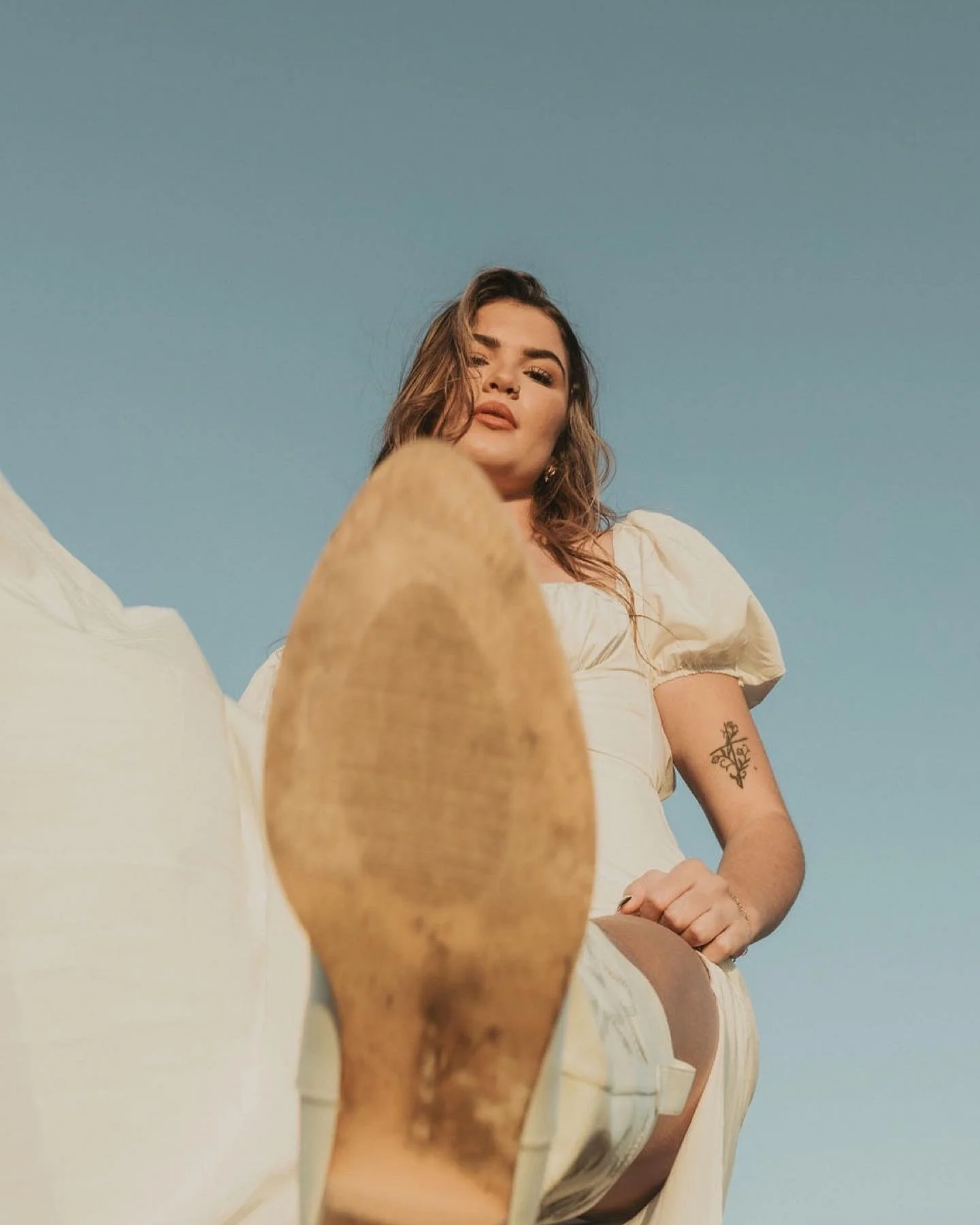 A woman with brown hair and tattoos on her left arm wearing a white dress outdoors against a clear blue sky.