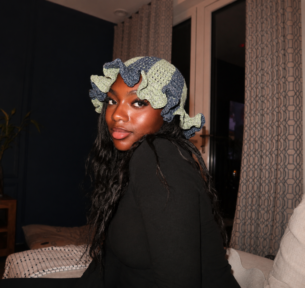 A woman with dark wavy hair wearing a blue and green patterned bonnet looks over her shoulder, sitting in a living room with patterned curtains and a dark wall.